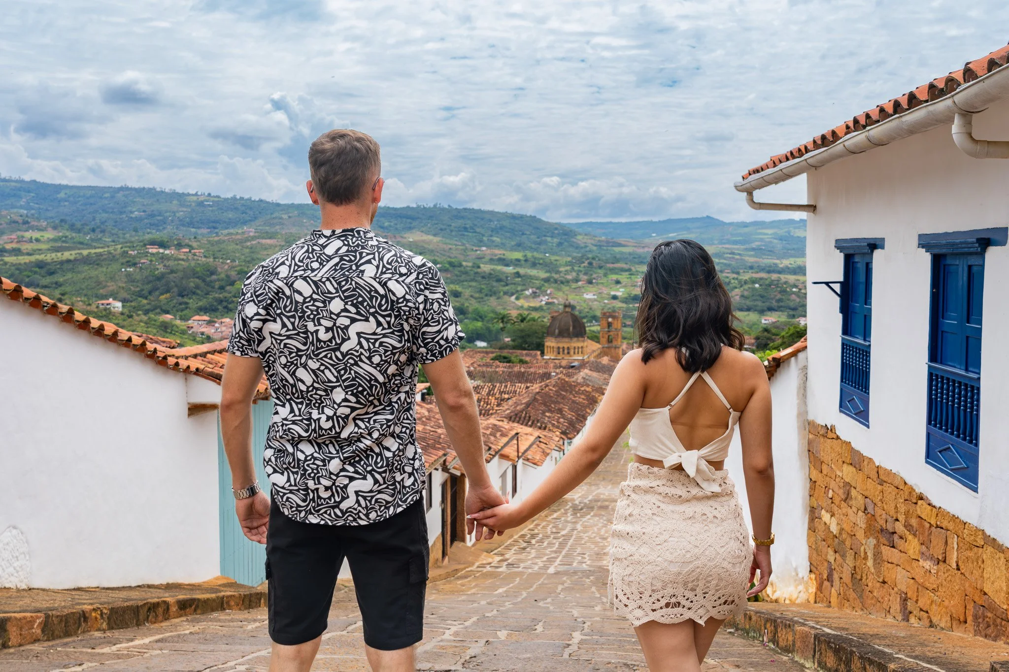 Pareja caminando de la mano por calle empedrada en un pueblo con casas blancas y puertas azules, rodeado de montañas verdes y cielo nublado.