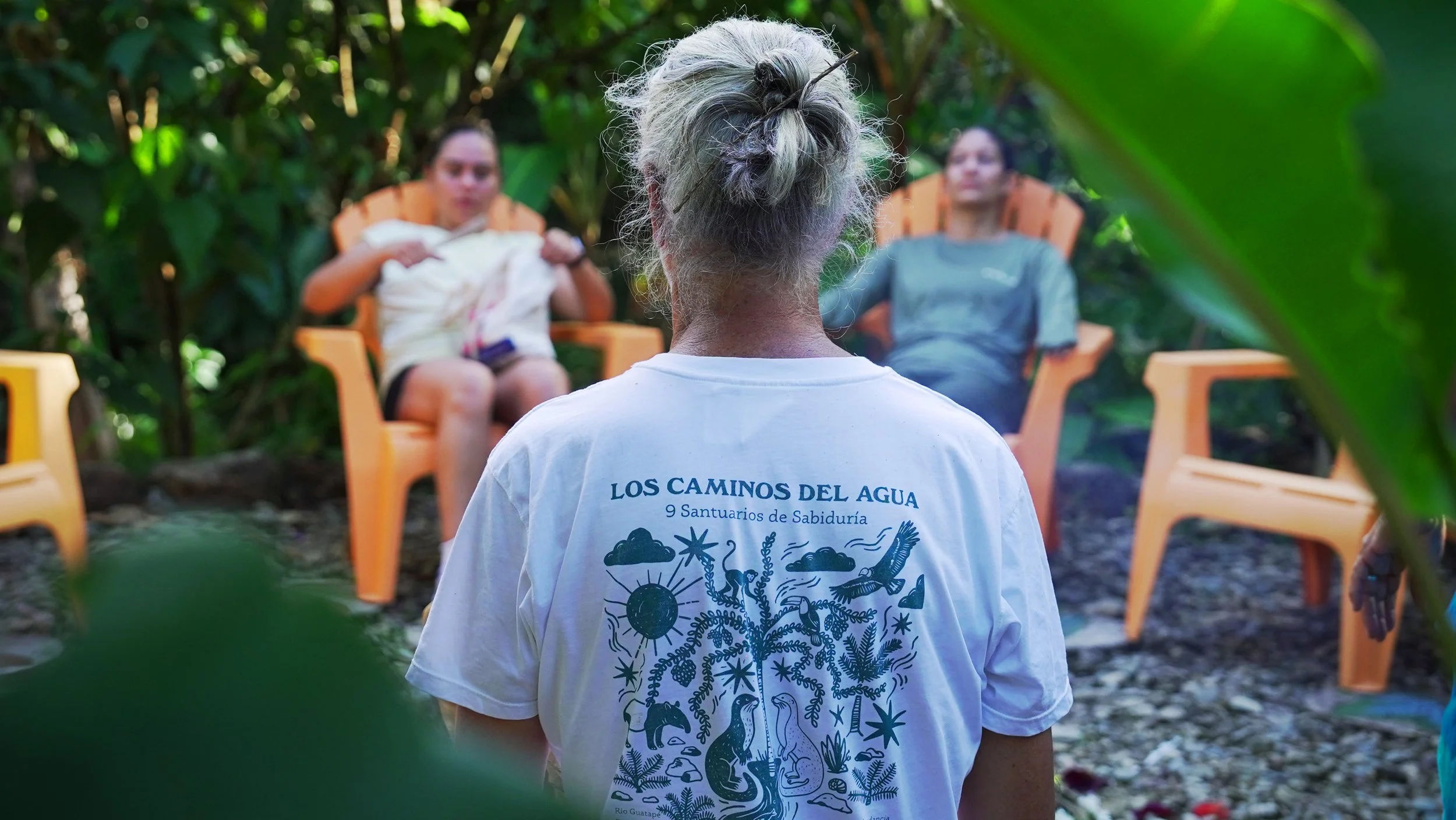 Persona viendo a dos personas recostadas en sillas de plástico en un entorno natural, con una especificación en la camiseta que dice 'LOS CAMINOS DEL AGUA' y '9 Santuarios de Sabiduría'