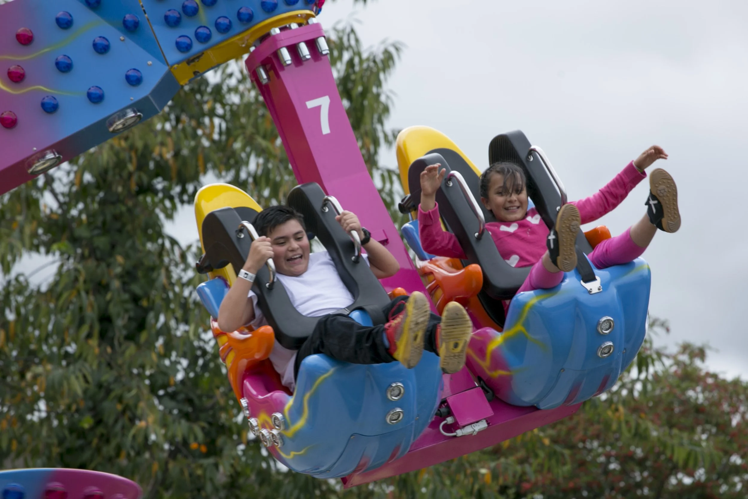 Dos niños sonriendo y disfrutando en una montaña rusa colorida en un parque de diversiones, con árboles de fondo.