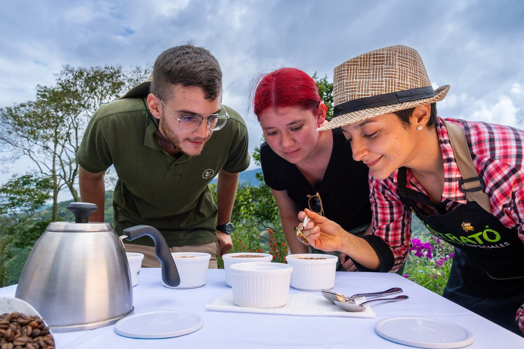 Tres personas observando diferentes muestras en tazas blancas en una mesa al aire libre, con árboles y cielo nublado de fondo.