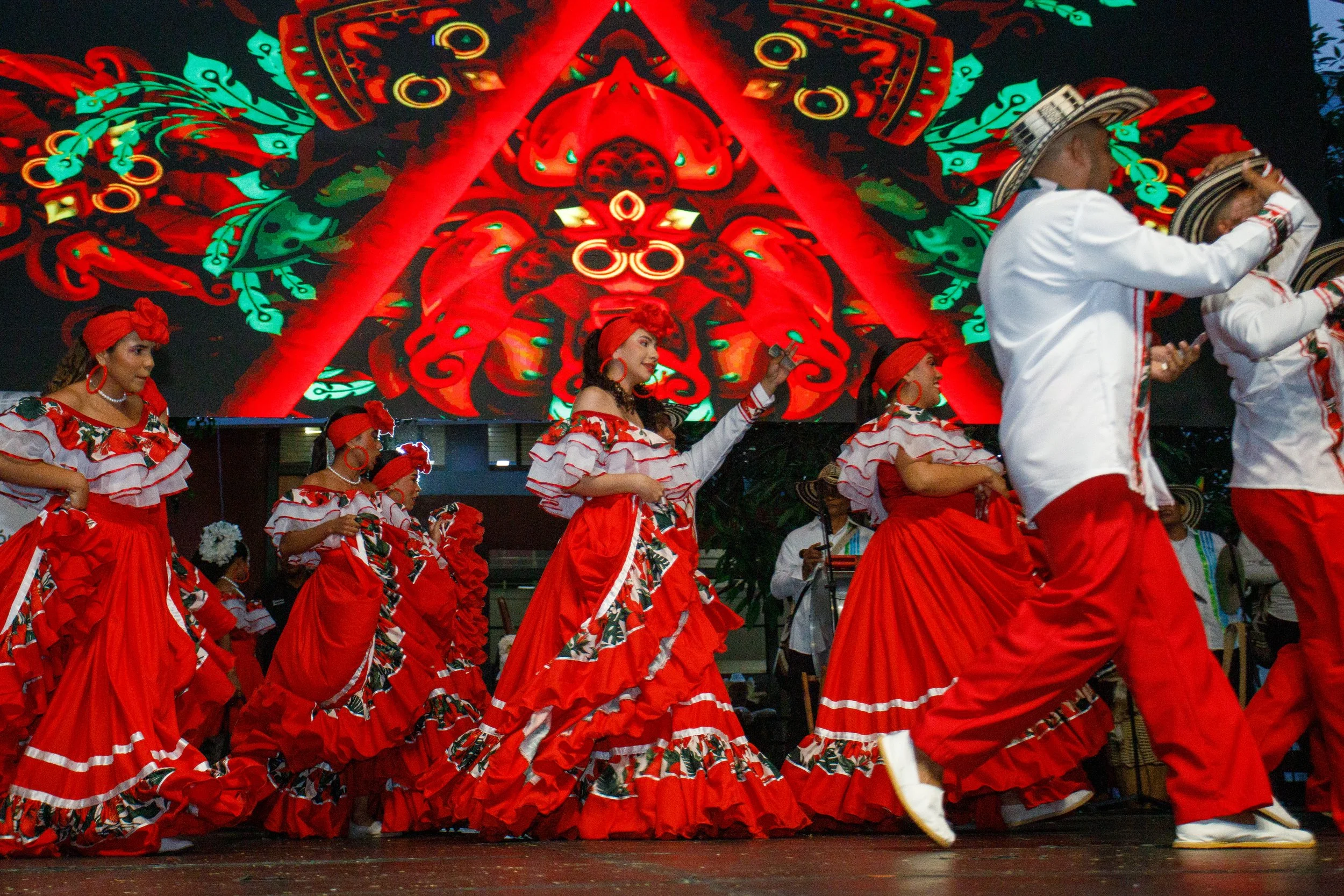 Grupo de mujeres bailando en un desfile con trajes tradicionales rojos con volantes blancos y motivos florales, acompañadas por músicos con trajes blancos y sombreros tradicionales, en un escenario con una pantalla grande que muestra un diseño colorido, posiblemente en una festival cultural.
