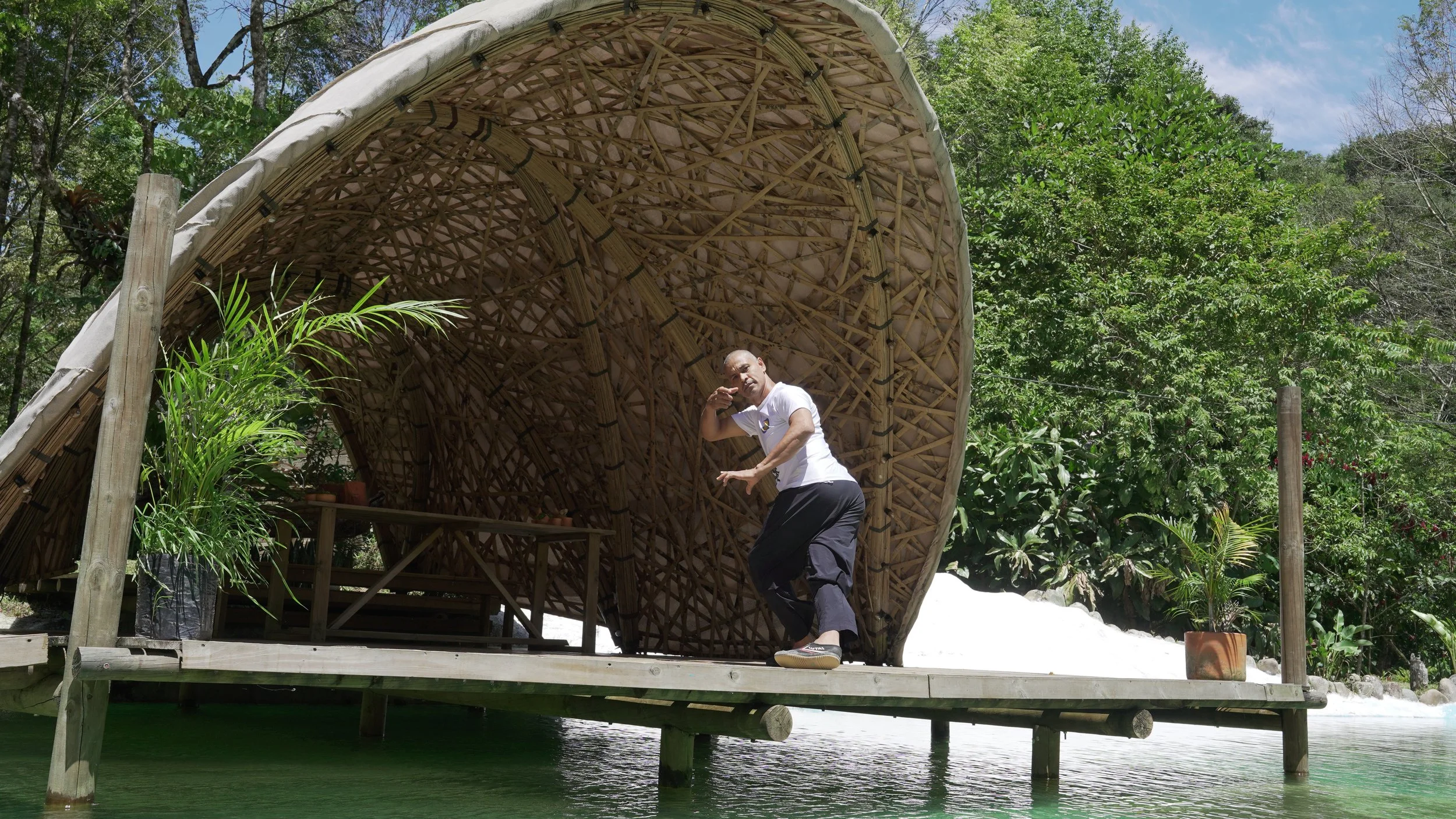 Hombre posando frente a una estructura de bambú en medio de la naturaleza, con plantas verdes y agua debajo