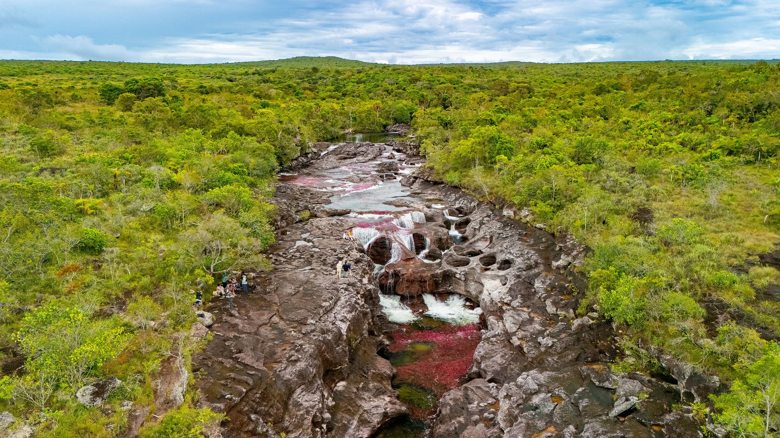 Paisaje natural con río rodeado de vegetación verde, montañas al fondo y cielo nublado.