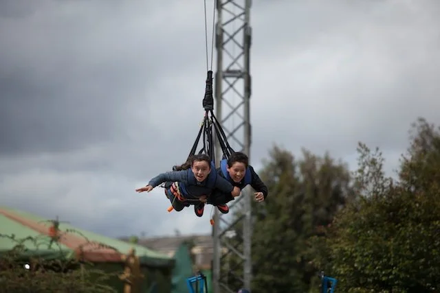 Dos personas en una atracción de parque de diversiones, colgadas en un asiento con cinturones de seguridad, parecen disfrutar y están en movimiento en el aire.