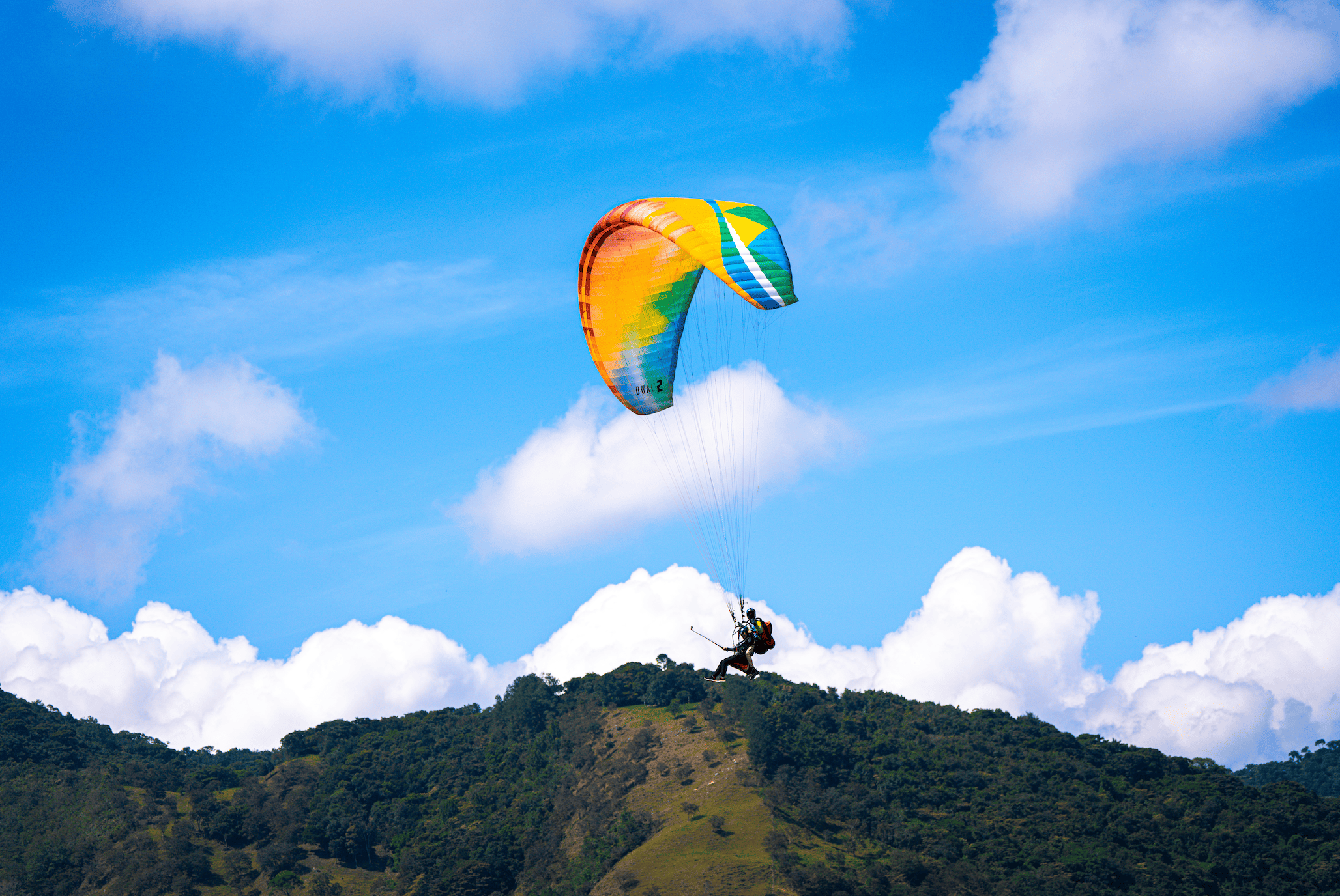 Persona practicando parapente en un cielo despejado con nubes y montañas verdes en el fondo.