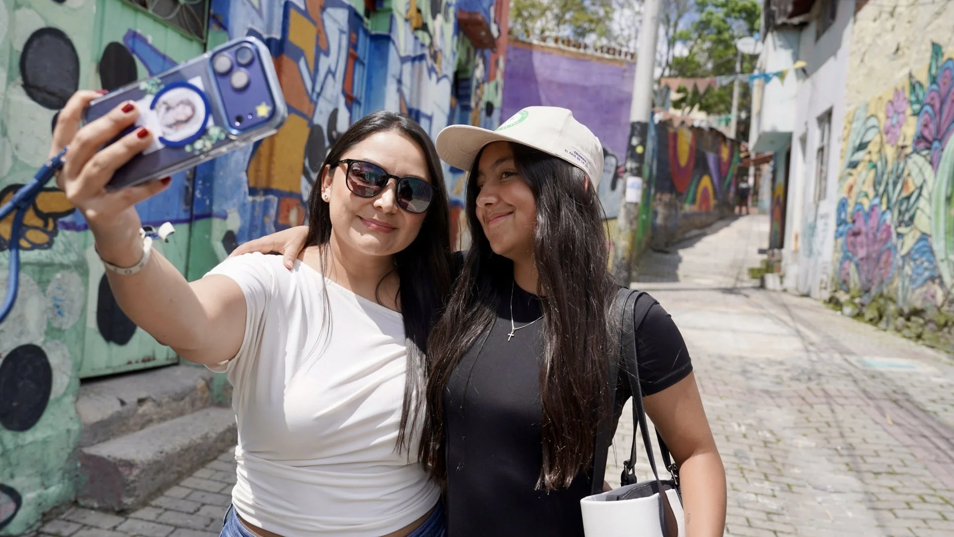 Dos mujeres toman una selfie en una calle con grafitis coloridos en las paredes.