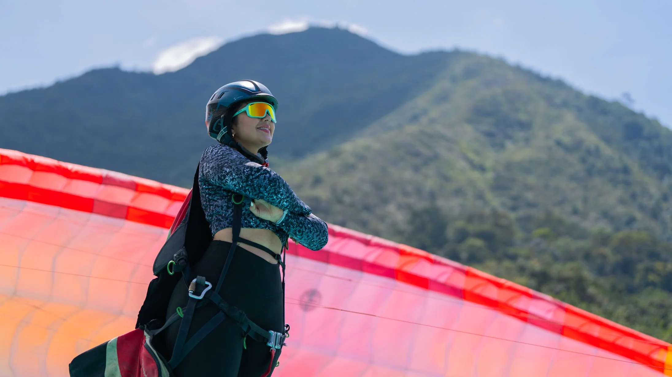 Mujer con equipo de parapente, gafas de sol, en un paisaje montañoso con cielo despejado.