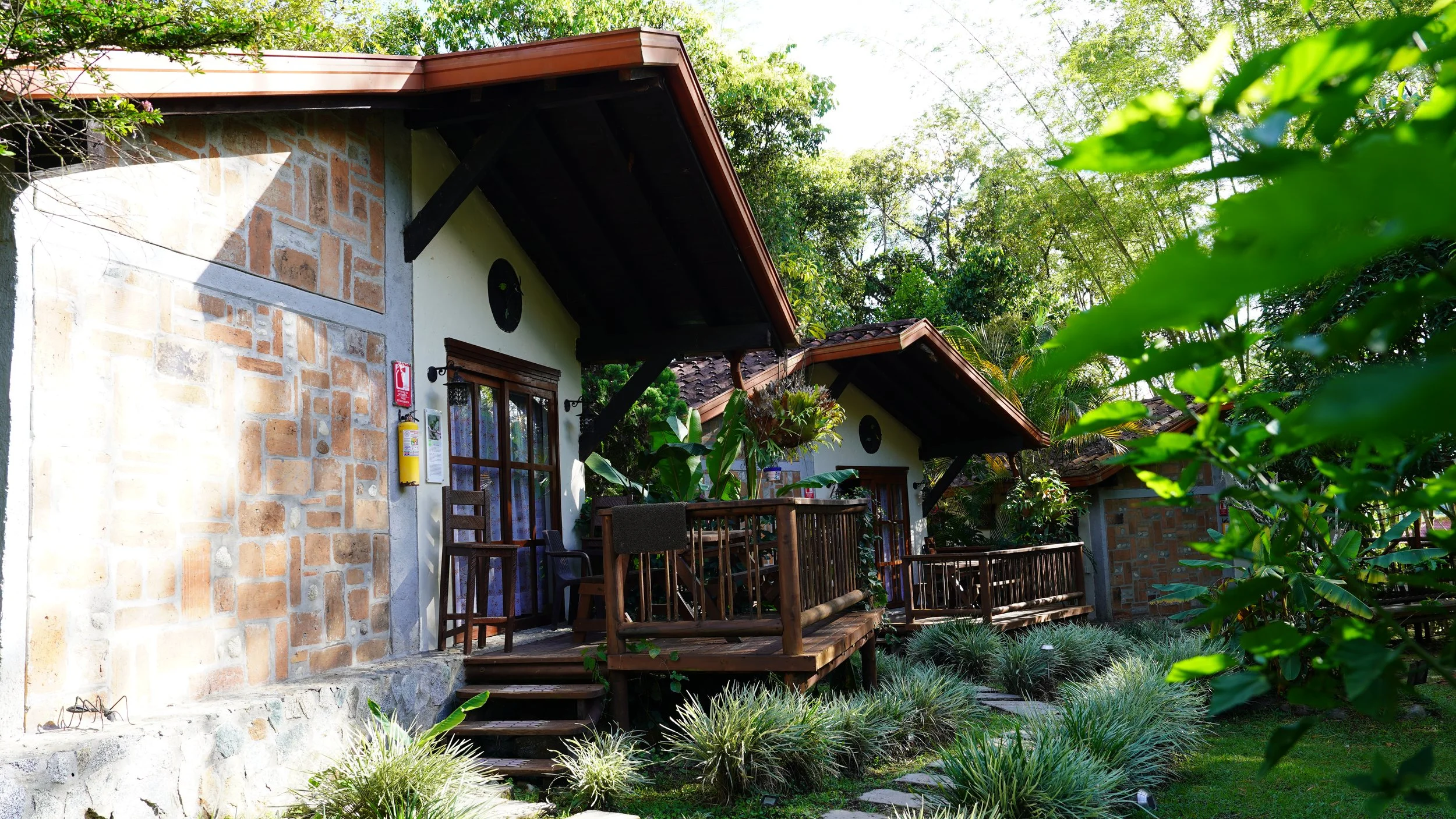 Casa de campo con paredes de ladrillo y ventanas de madera, rodeada de vegetación verde y un balcón de madera.