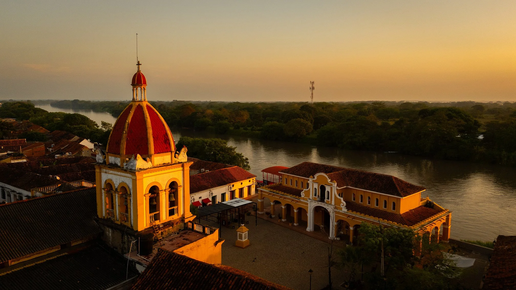 Vista de una iglesia con cúpula roja y torre, en una ciudad junto a un río rodeado de árboles, al atardecer con cielo dorado.