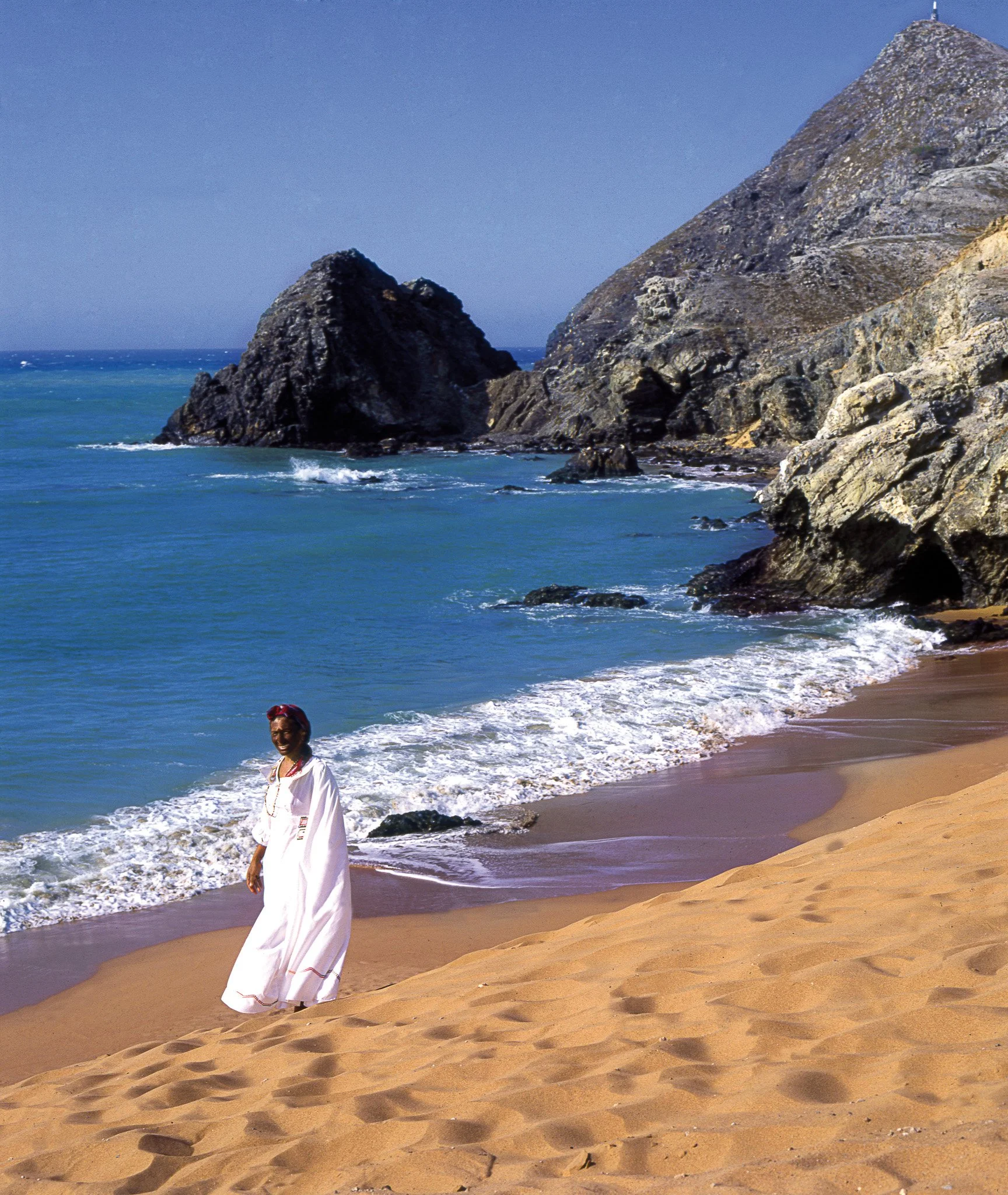 Mujer caminando por la playa con olas y rocas en el fondo, vestido blanco y sombrero rojo.