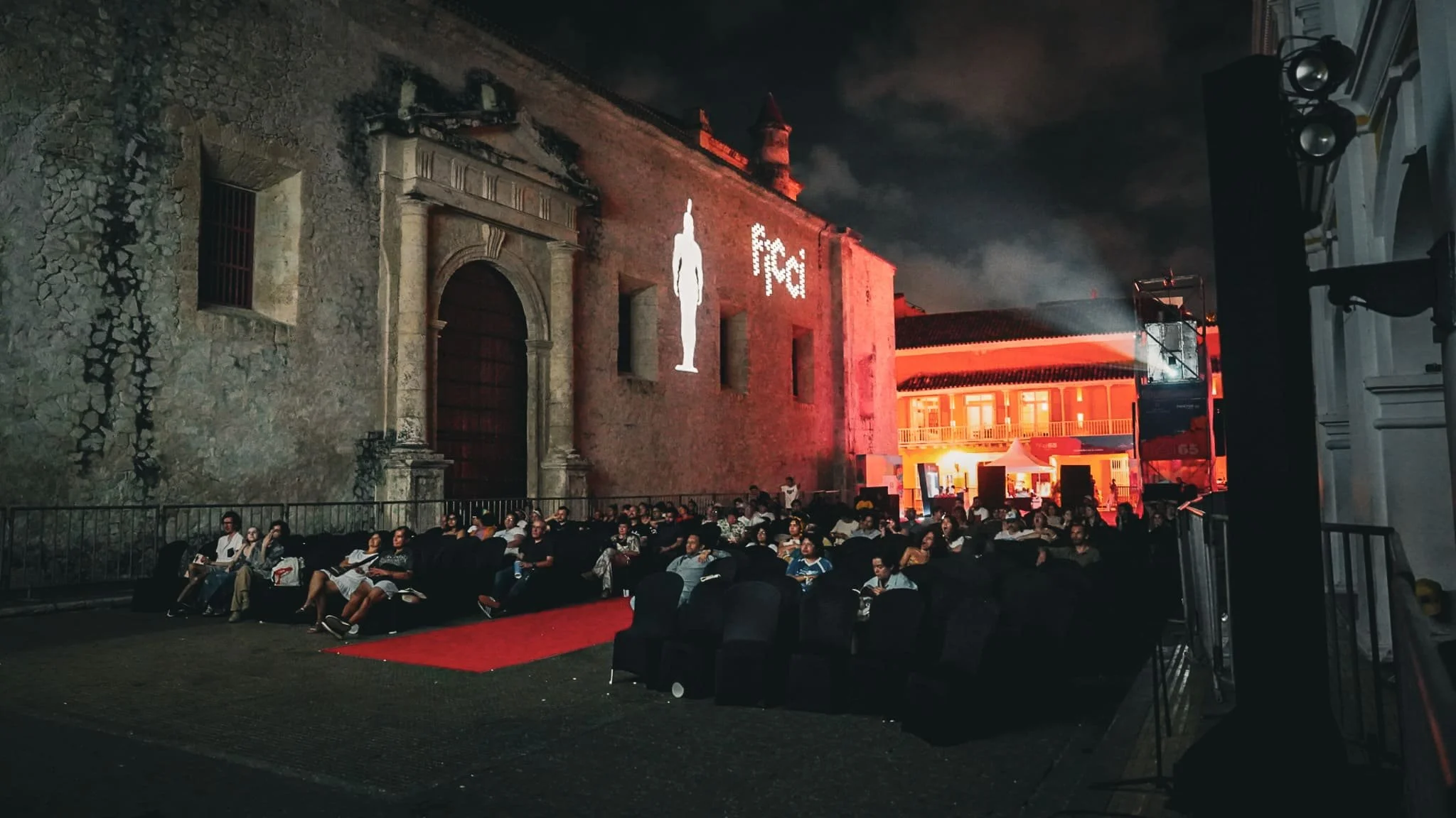 Audiencia al aire libre en un casco antiguo iluminado, con muchas personas sentadas, un escenario en el fondo con luces y una pantalla gigante con la palabra 'fat'.