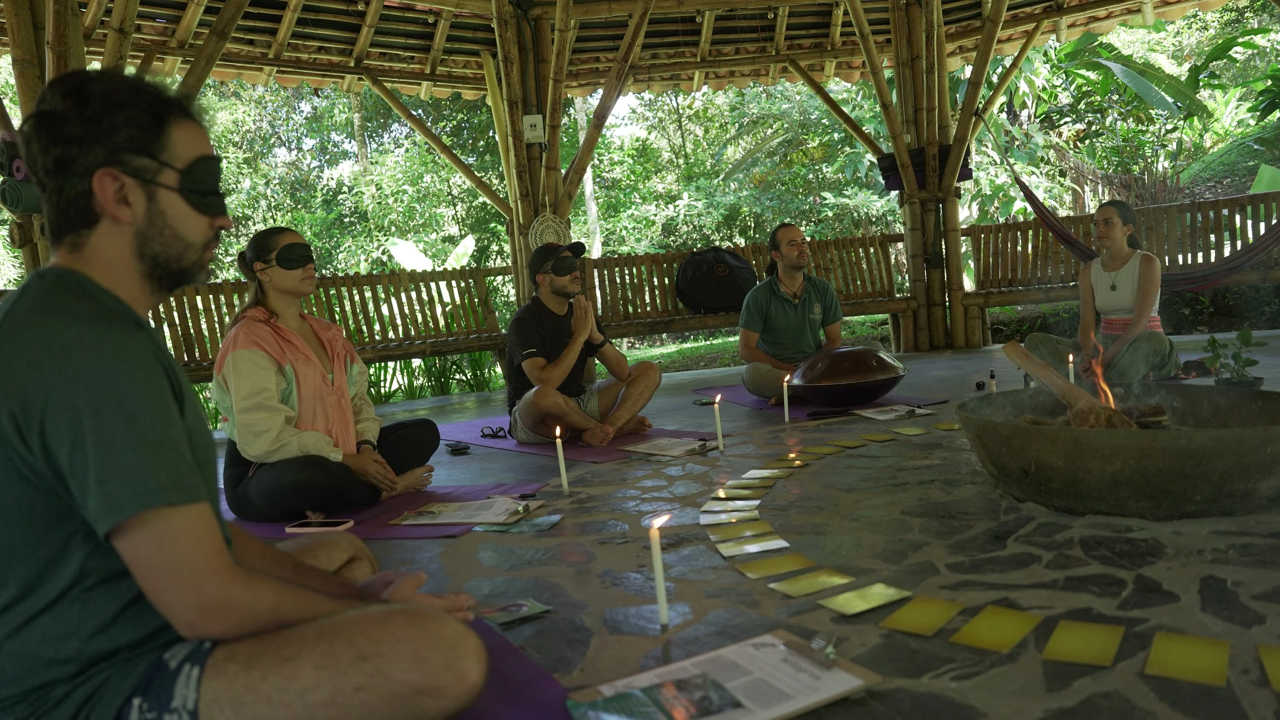 Grupo de personas participando en una ceremonia espiritual en un espacio abierto con estructura de madera, rodeados de naturaleza, algunas con ojos vendados, en postura de meditación y oración, con velas y objetos sagrados en el centro.
