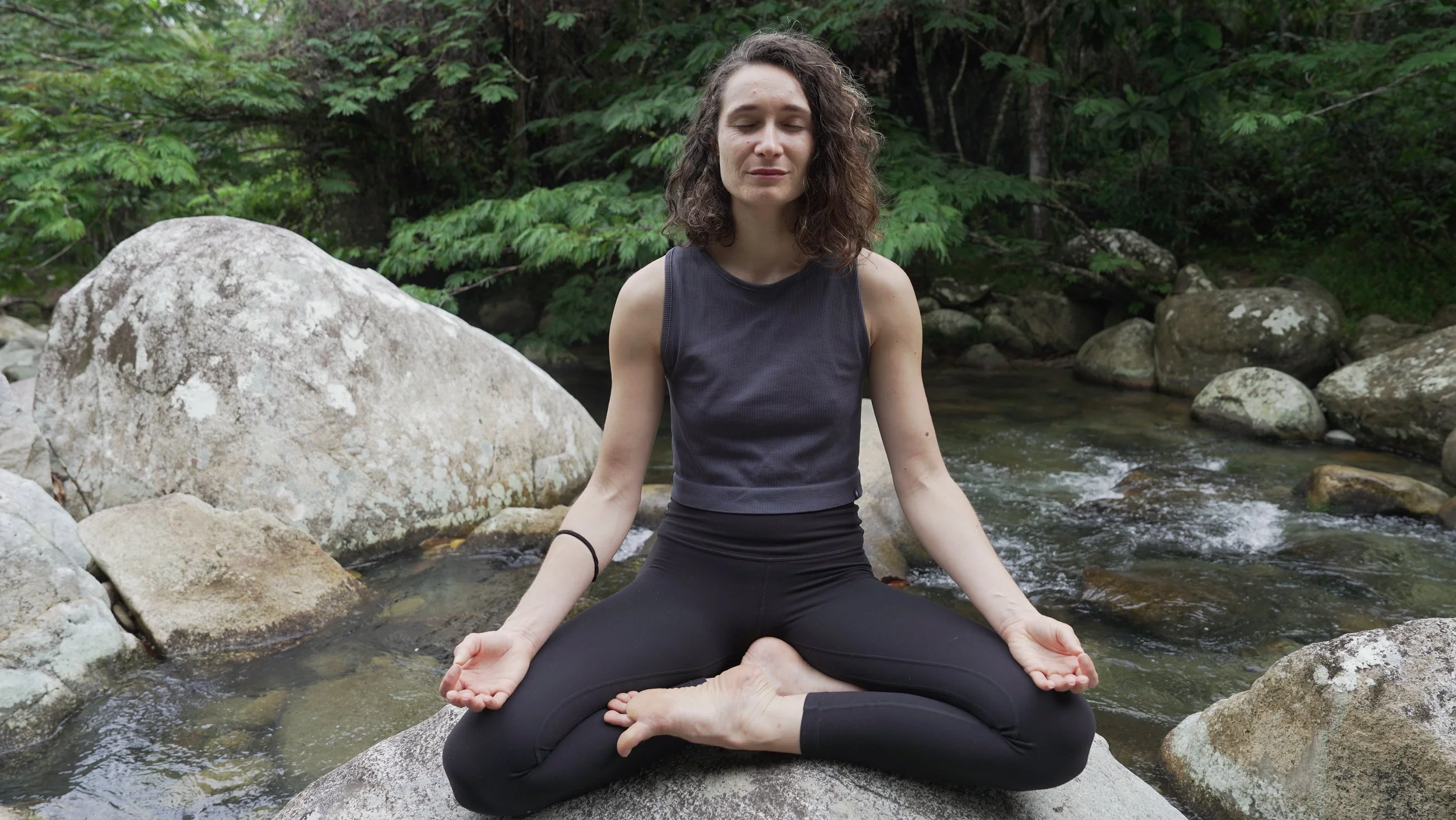 Mujer practicando yoga en una roca junto a un río en un entorno natural con árboles verdes.