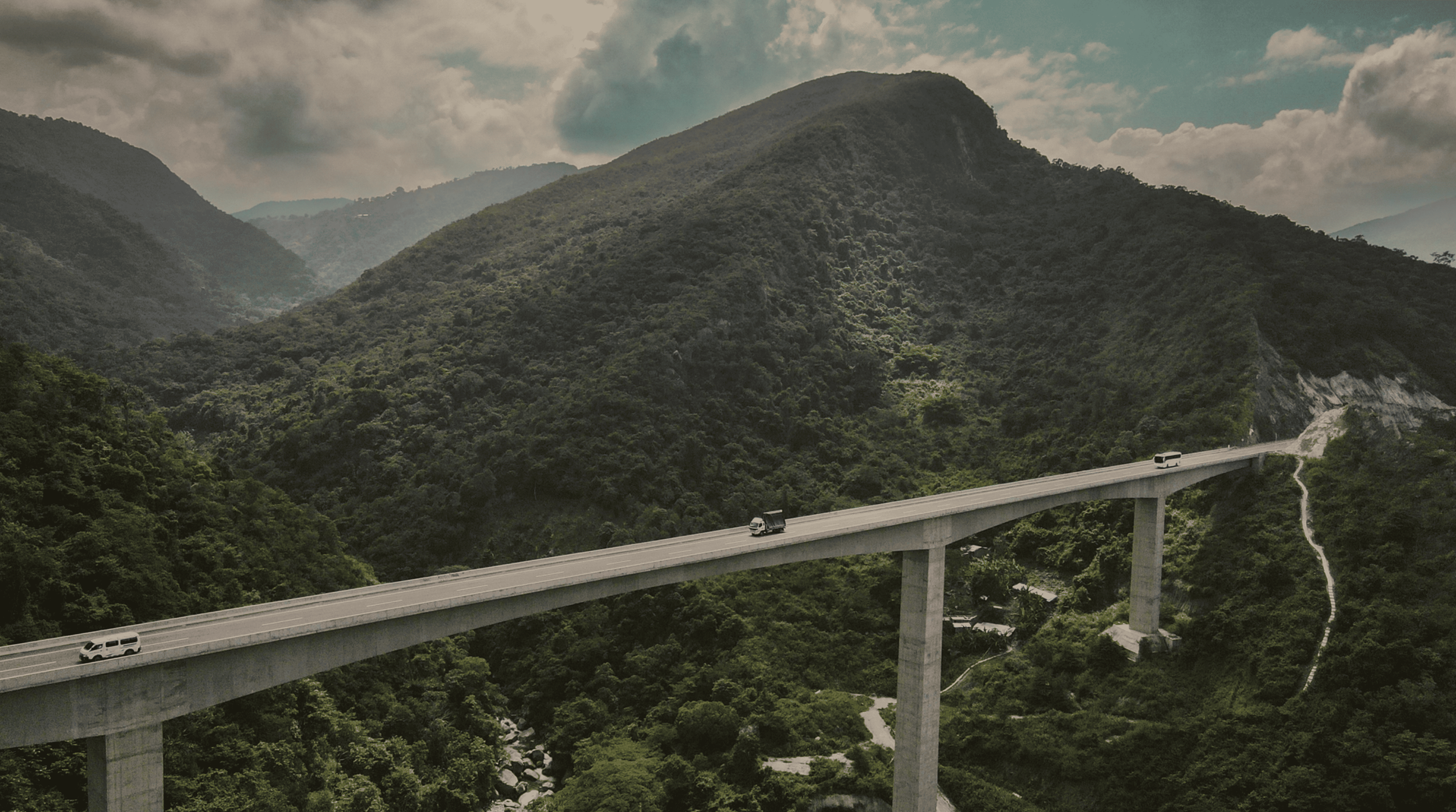 Puente elevado en medio de montañas verdes con caminos y vehículos transitando, cielo nublado