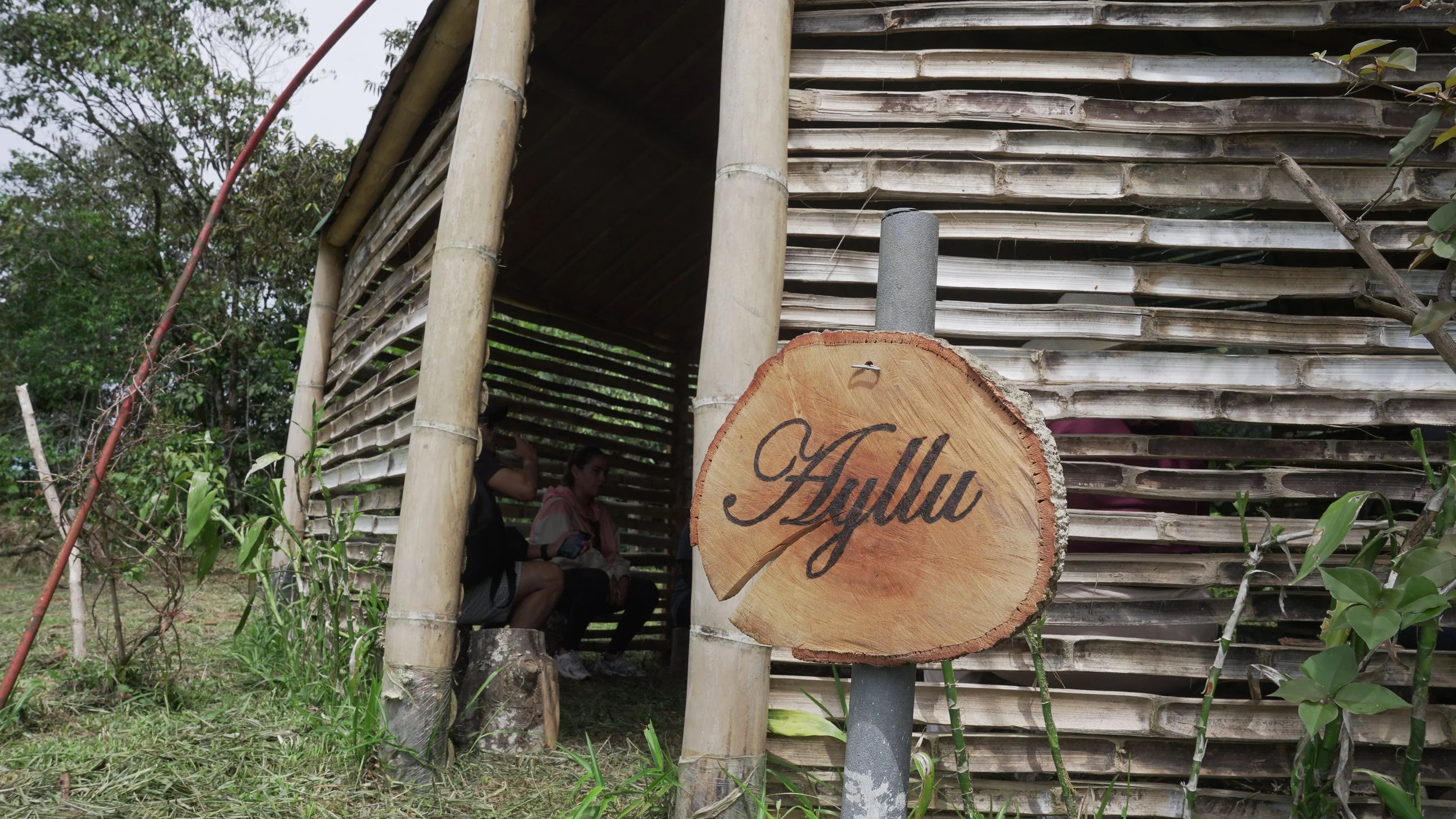 Estructura de madera con un cartel que dice 'Ayllu', rodeada de vegetación y dos personas sentado adentro.