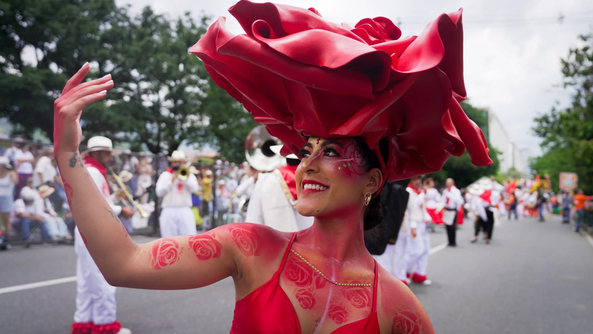 Mujer vestida con un vestido rojo y un sombrero grande de tela en forma de rosa durante un desfile, rodeada de otras personas y músicos en la calle.