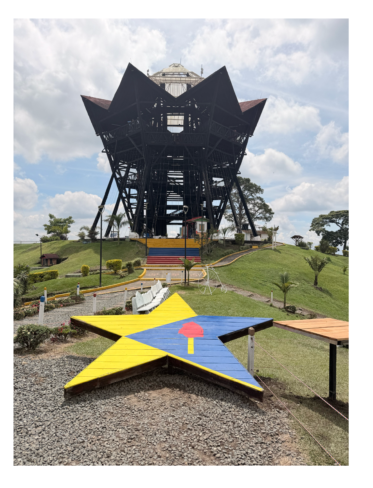 Una torre de observación de estructura metálica con forma de flor y una pasarela en forma de estrella con colores amarillo, azul y rojo, en un parque con césped, árboles y caminos pavimentados.