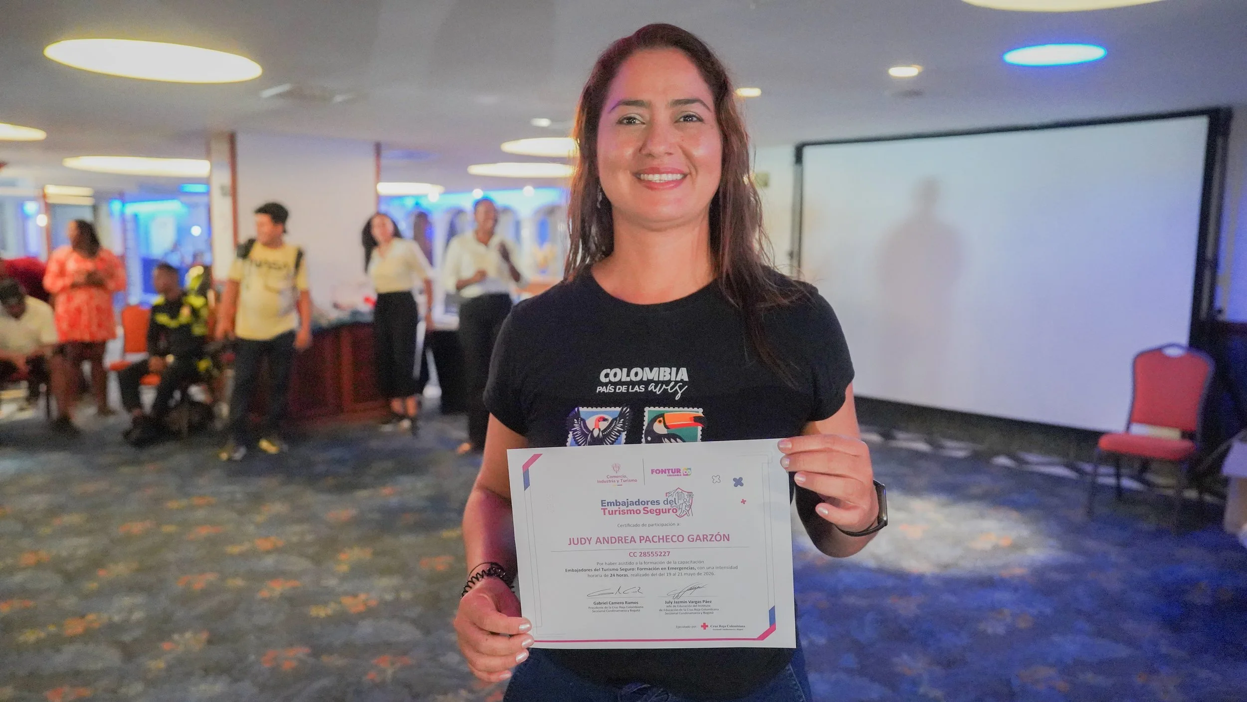Mujer sosteniendo un certificado en un evento, con otras personas en el fondo en un lugar cerrado.