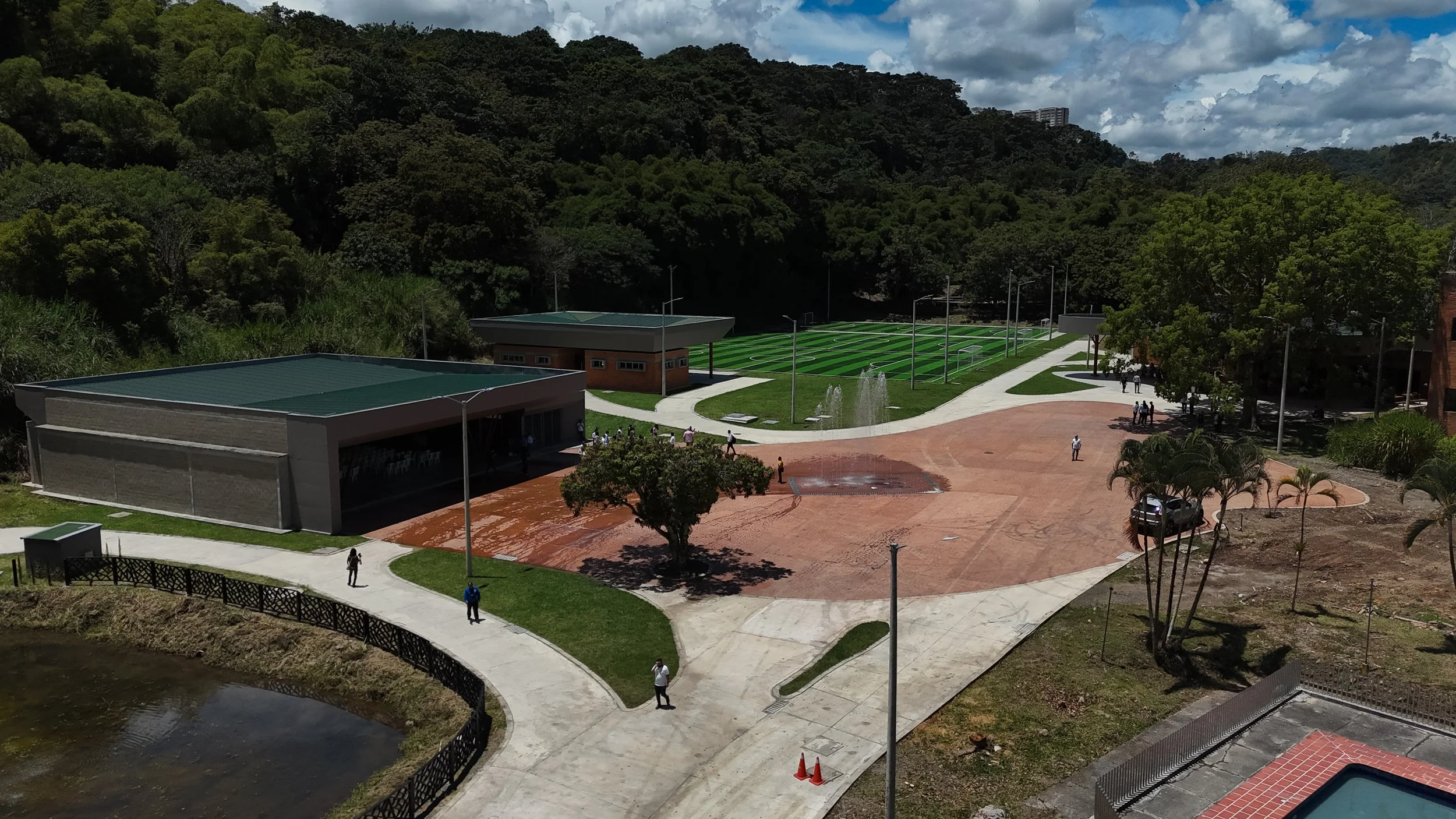 Vista aérea de una cancha de fútbol y una pista de atletismo en un parque rodeado de árboles verdes y un cielo nublado.
