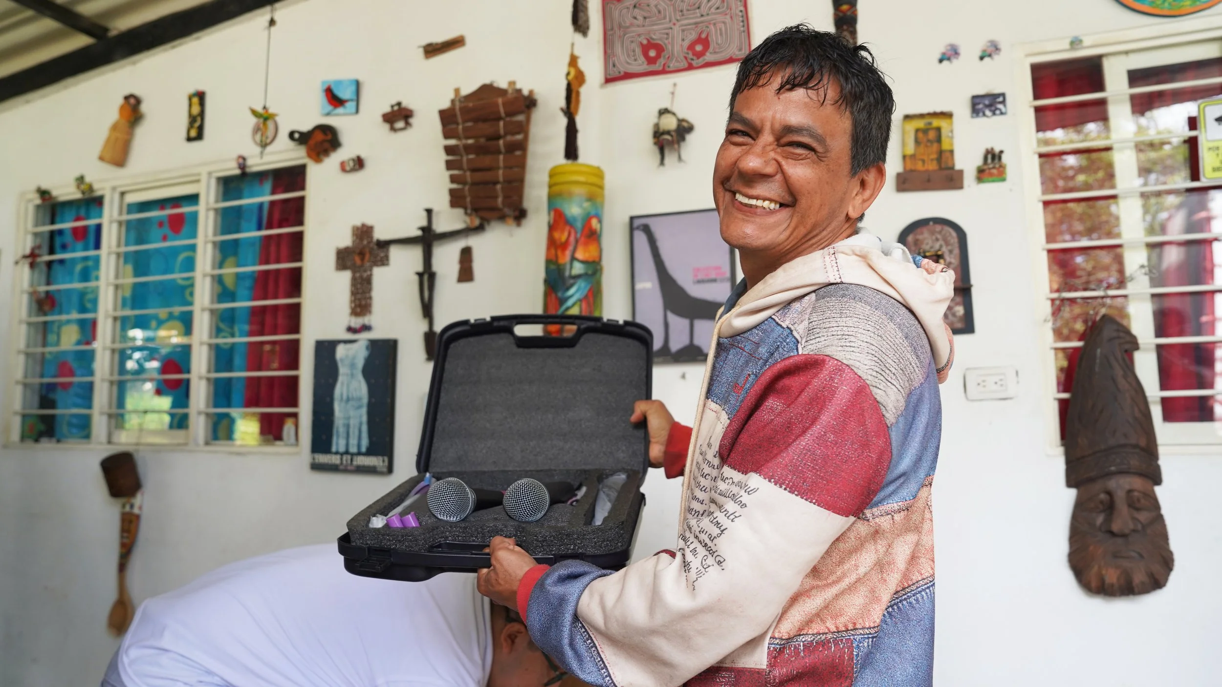 Hombre sonriendo sosteniendo una caja con micrófonos en un ambiente decorado con arte y objetos culturales.