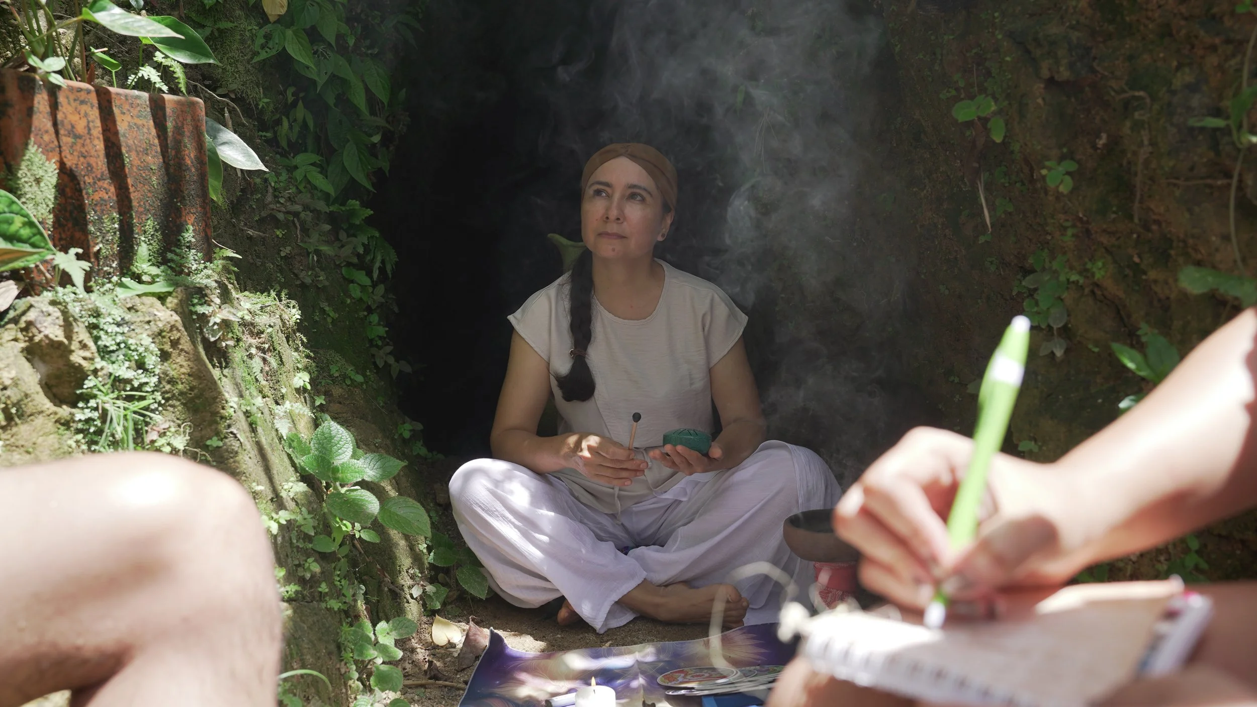 Mujer sentada en una cueva o un hueco en la tierra, rodeada de plantas verdes, con humo o vapor en el aire, sosteniendo un dispositivo y con un hombre en el primer plano usando un bolígrafo y un cuaderno.