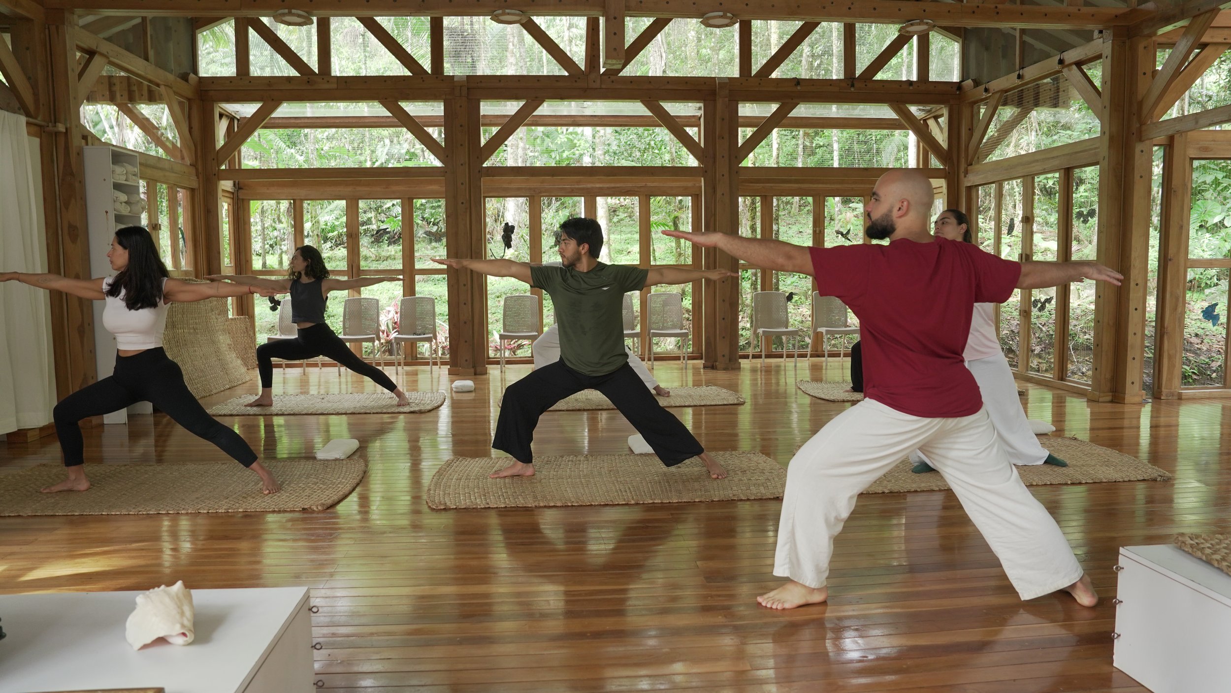 Grupo de personas practicando yoga en un salón de madera rodeado de naturaleza.