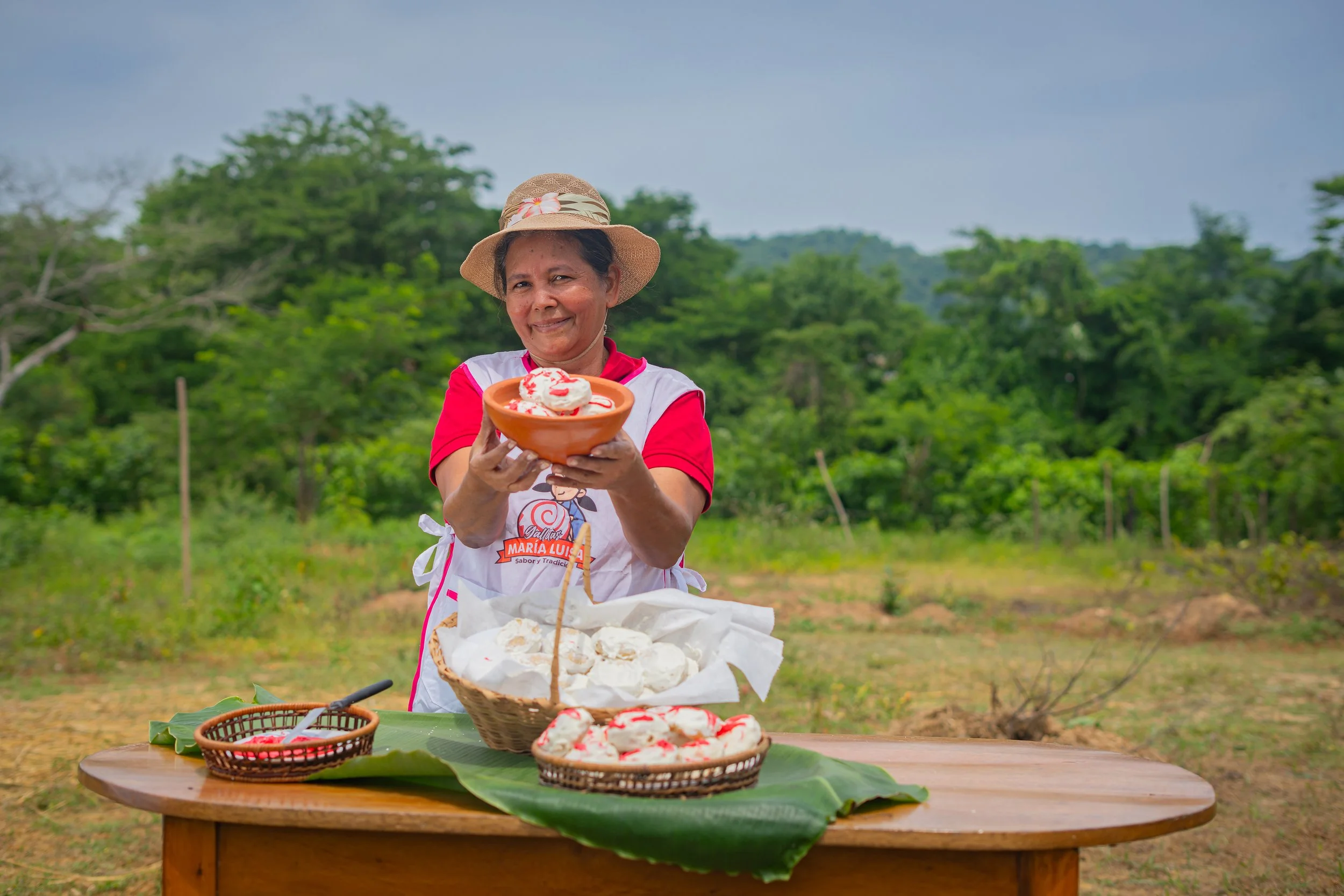 Mujer en campo rural sosteniendo un plato con postre de helado o nieve, con varias mesas y cestas con pan o productos similares en primer plano.