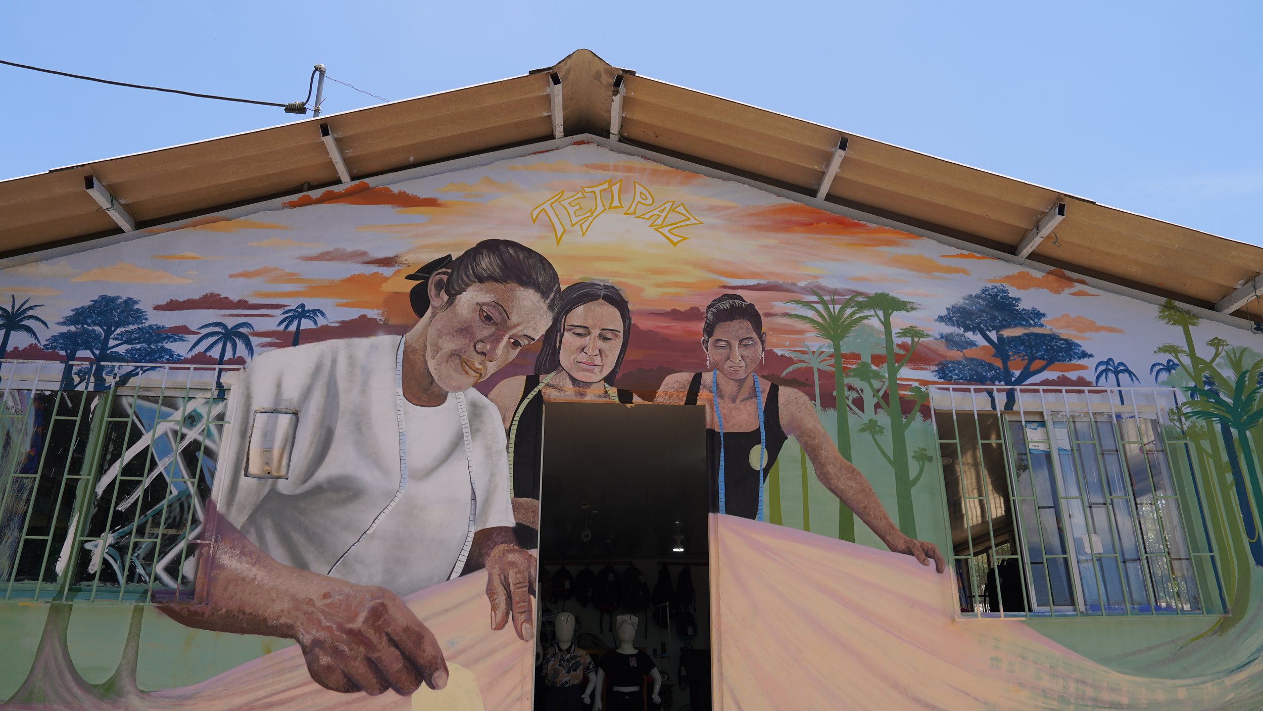 Mural colorido que representa a tres mujeres trabajando juntas en un entorno natural, con árboles y un cielo al atardecer, y la palabra 'Turipaz' en la parte superior.