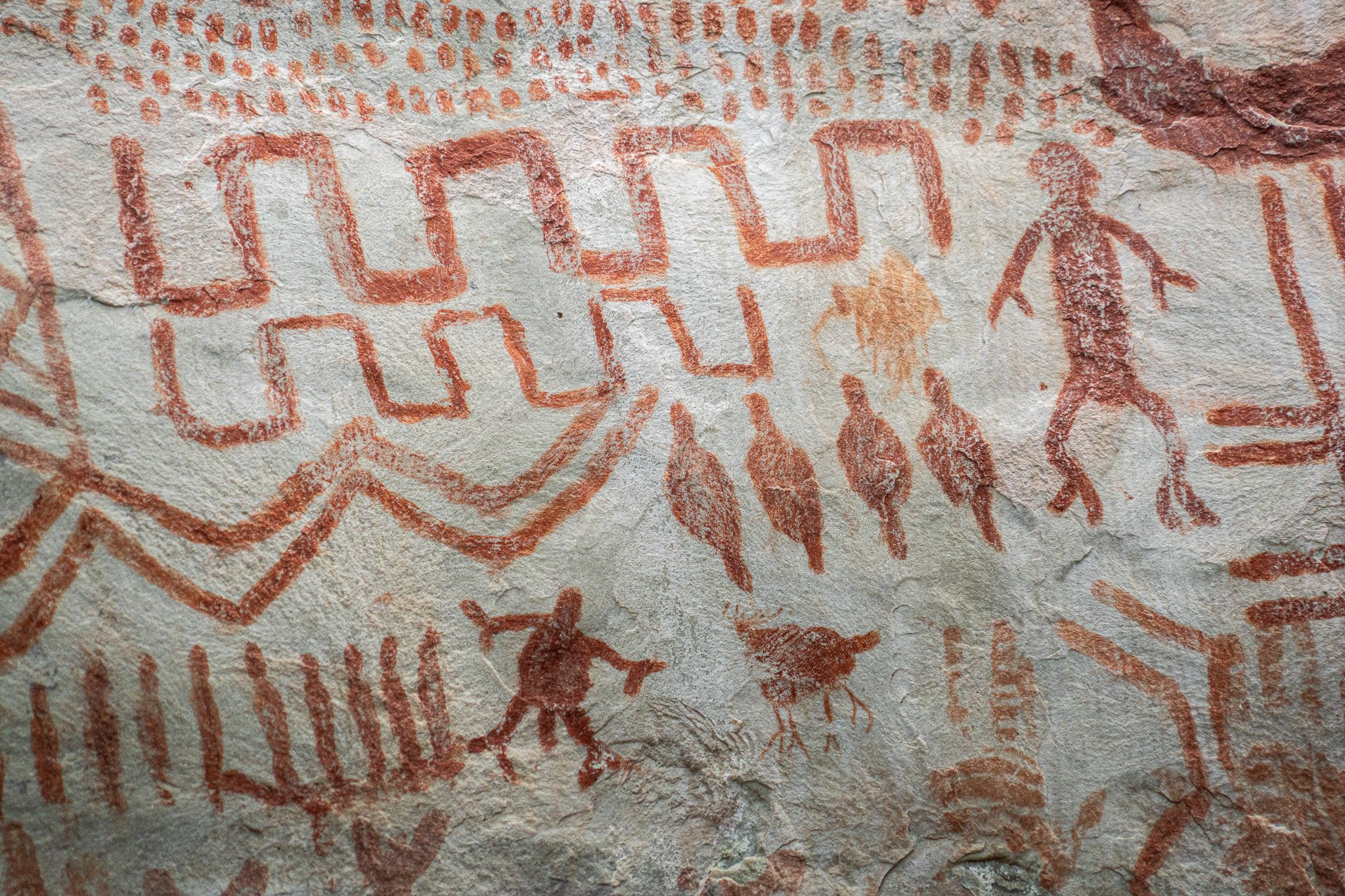 Arte rupestre indígena con figuras humanas y animales en una pared de piedra, pintadas en color rojo y ocres.