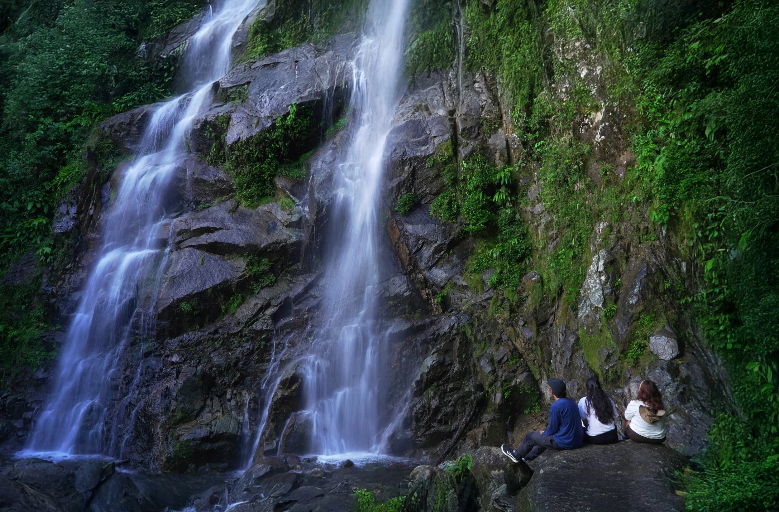 Vista de una cascada en medio de una selva con personas observando desde las rocas.