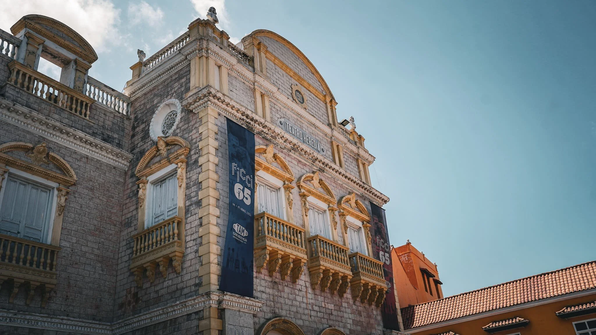 Edificio antiguo de ladrillos con balcones y decoraciones en blanco y dorado, cartel que promociona una exposición en el teatro, y un cielo despejado en el fondo.