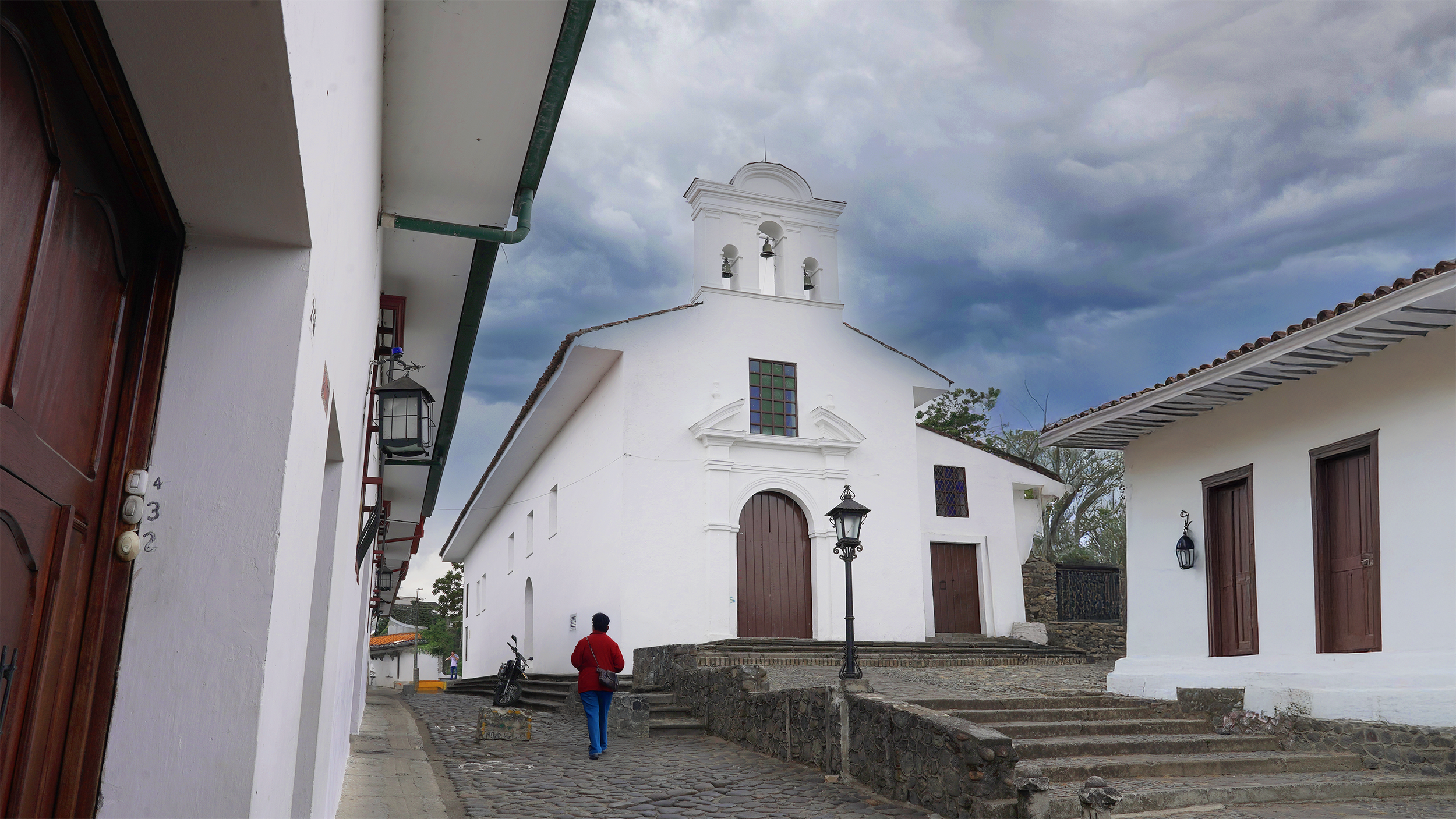 Plaza con iglesia blanca y campanario, calles empedradas, faroles antiguos y cielo nublado.