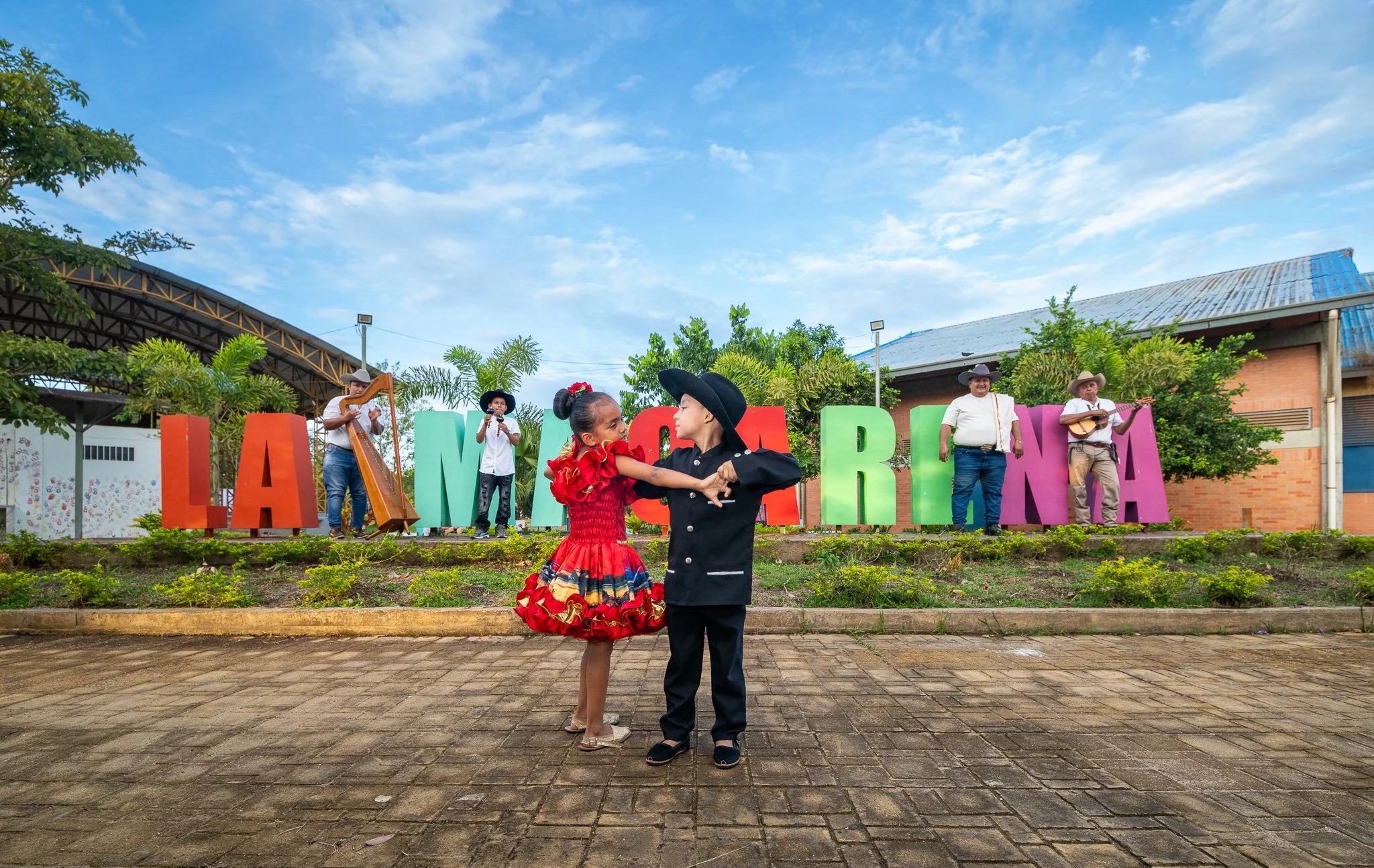Pareja bailando en una fiesta tradicional con músicos tocando en el fondo, letras grandes de colores que dicen 'LA MIERDA' y vegetación alrededor.