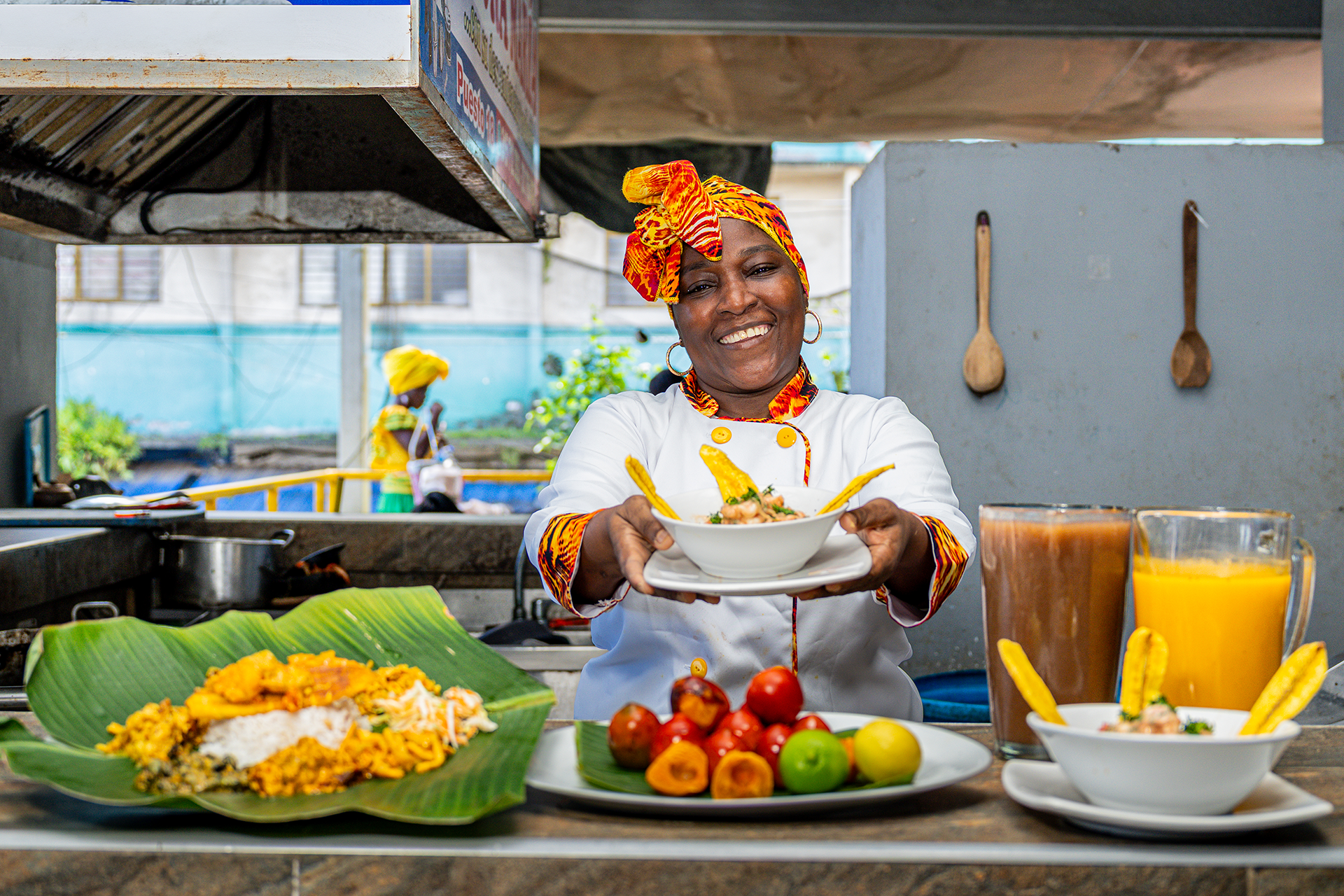 Mujer sonriente con banda en la cabeza, entregando un plato de comida en un puesto de comida callejera, con diversos platillos y frutas en la mesa delante suyo.