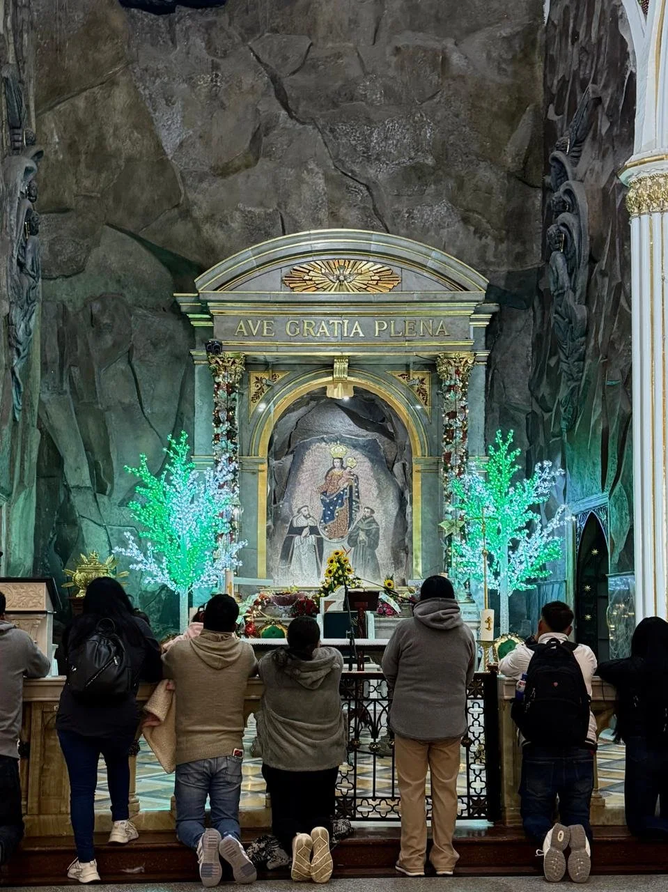 Personas rezando frente a un altar en una cueva, con decoraciones navideñas y un mosaico religioso en el fondo.