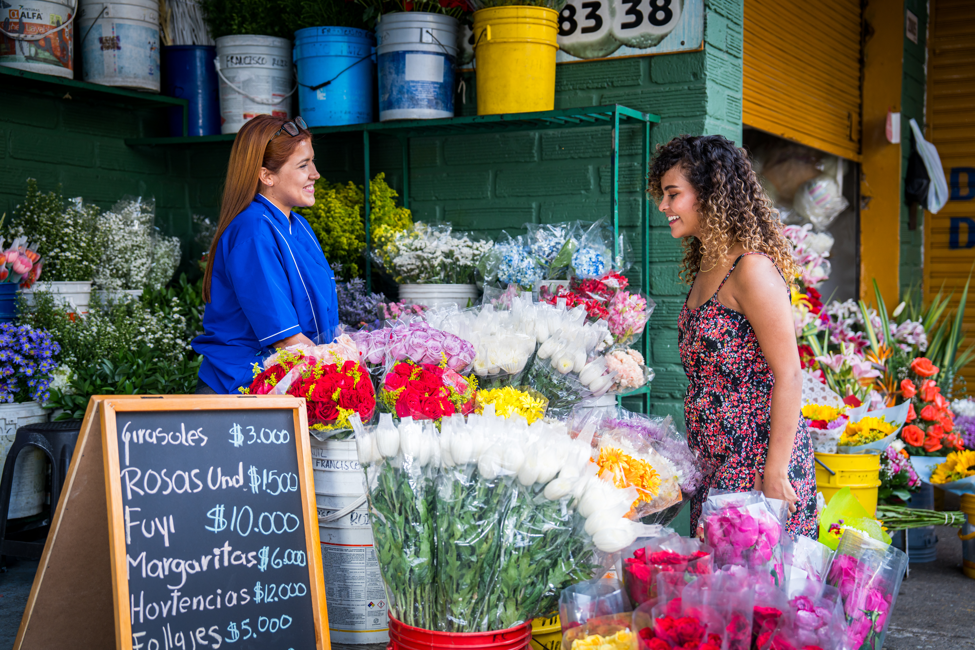 Mujer comprando flores en un puesto de mercado, atendido por una vendedora con uniforme azul. Hay muchas flores de colores y un cartel con precios escritos en español.