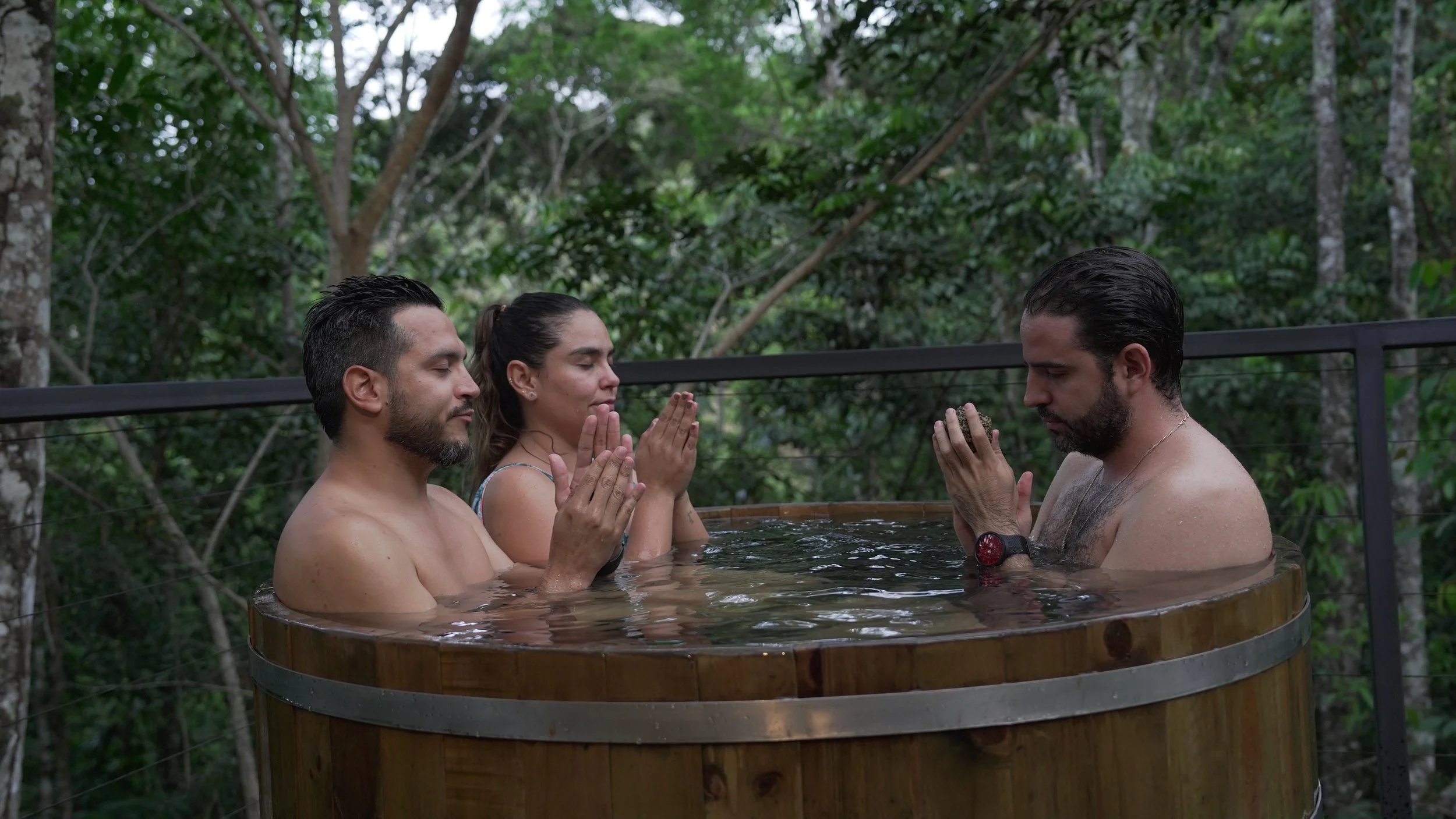 Cuatro personas, dos hombres y una mujer, en un jacuzzi en medio de la naturaleza, con ojos cerrados y manos juntas en oración o meditación.
