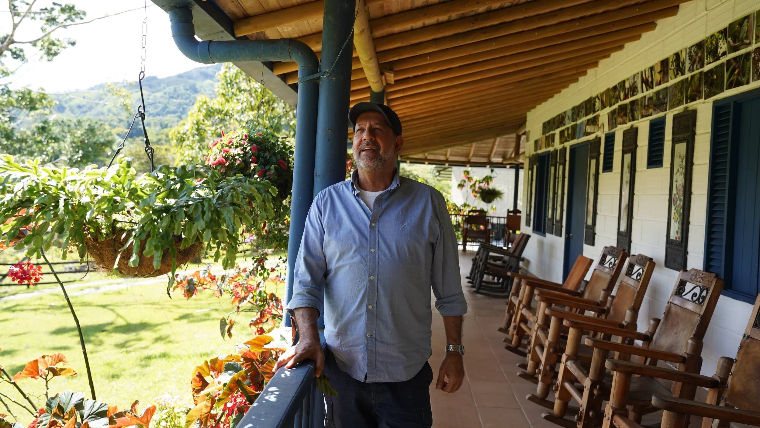 Un hombre con camisa de manga larga en un porche con sillas de madera, plantas colgantes y un paisaje de montañas al fondo.