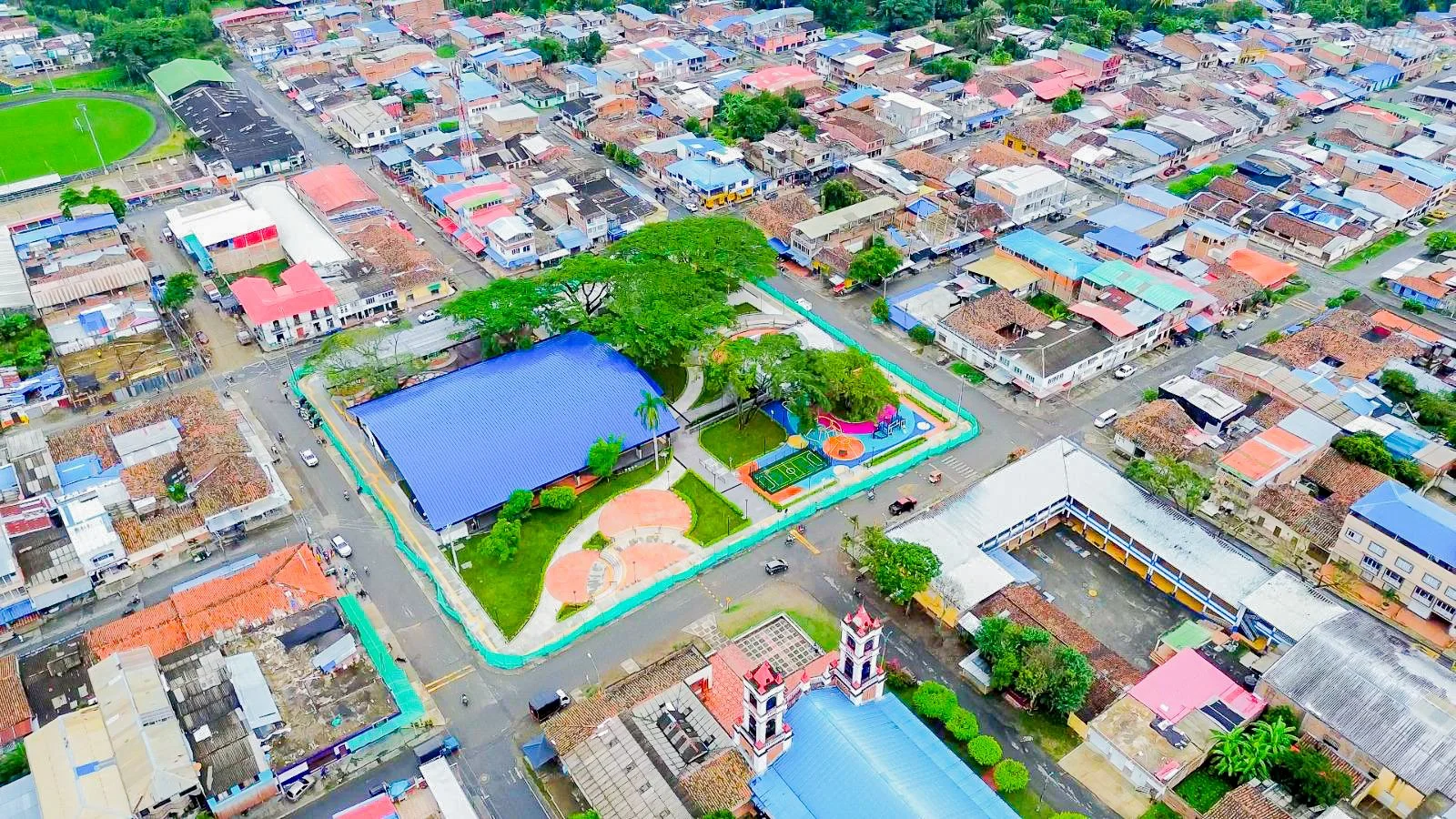 Vista aérea de un vecindario con una escuela que tiene un techado azul y un área de juegos, rodeada de casas con techos de diferentes colores. Incluye un campo de fútbol y un parque con árboles y áreas de recreo.