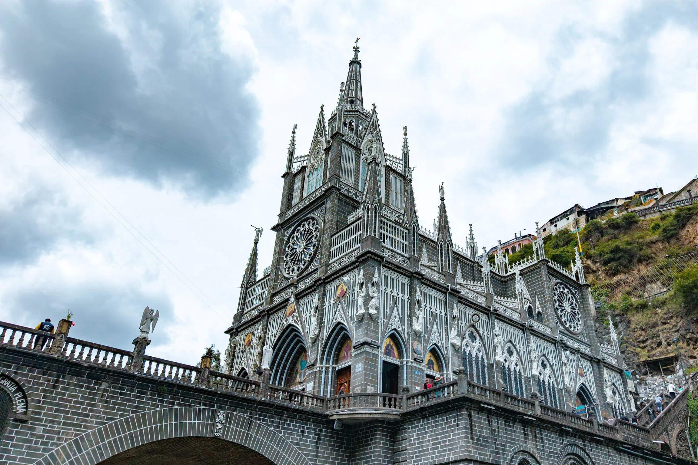 Iglesia de Las Lajas en Pasto, Nariño, de color gris oscuro con detalles en blanco y vitrales, ubicada en una ladera con casas alrededor, bajo un cielo nublado.