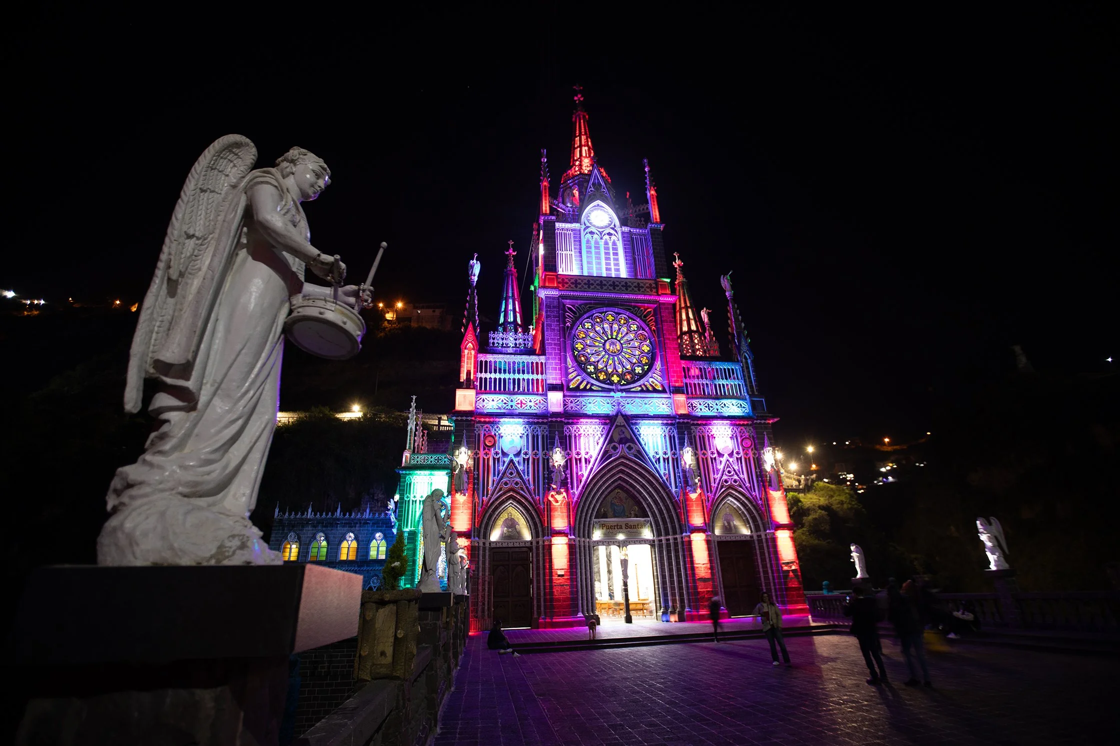 Catedral de coloridas luces iluminada de noche, con una estatua de un ángel en primer plano a la izquierda.