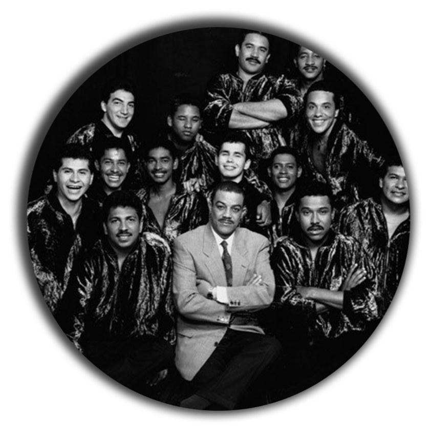 Grupo de personas en vestimenta de escenario, algunos con uniformes de escenario brillantes, y un hombre en el centro con traje gris y expresión seria, en una fotografía en blanco y negro.