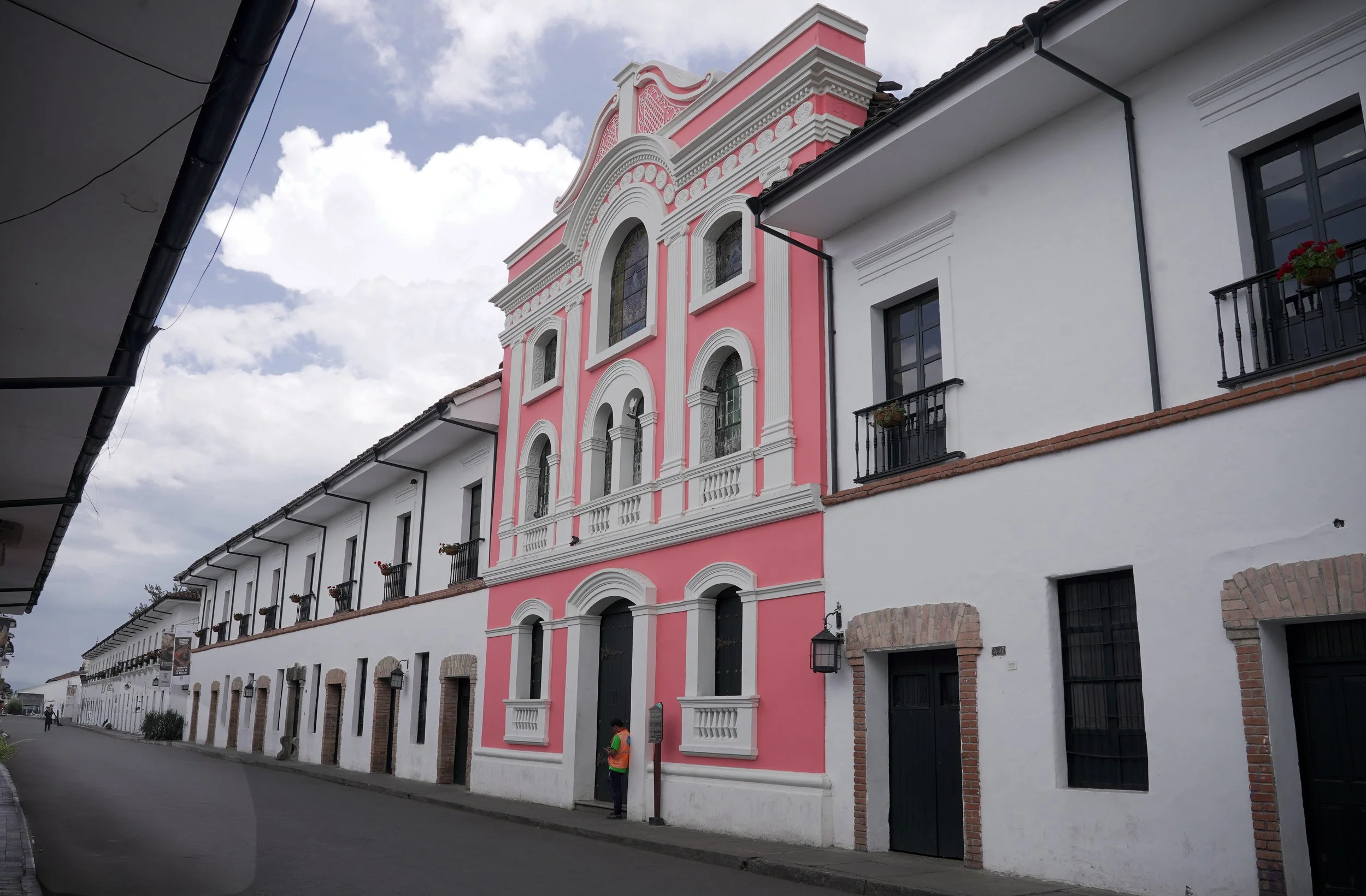 Edificio pintado de color rosa y blanco con estilo colonial, con ventanas de arcos y detalles decorativos, en una calle con edificaciones blancas y balcones con flores, bajo un cielo nublado.