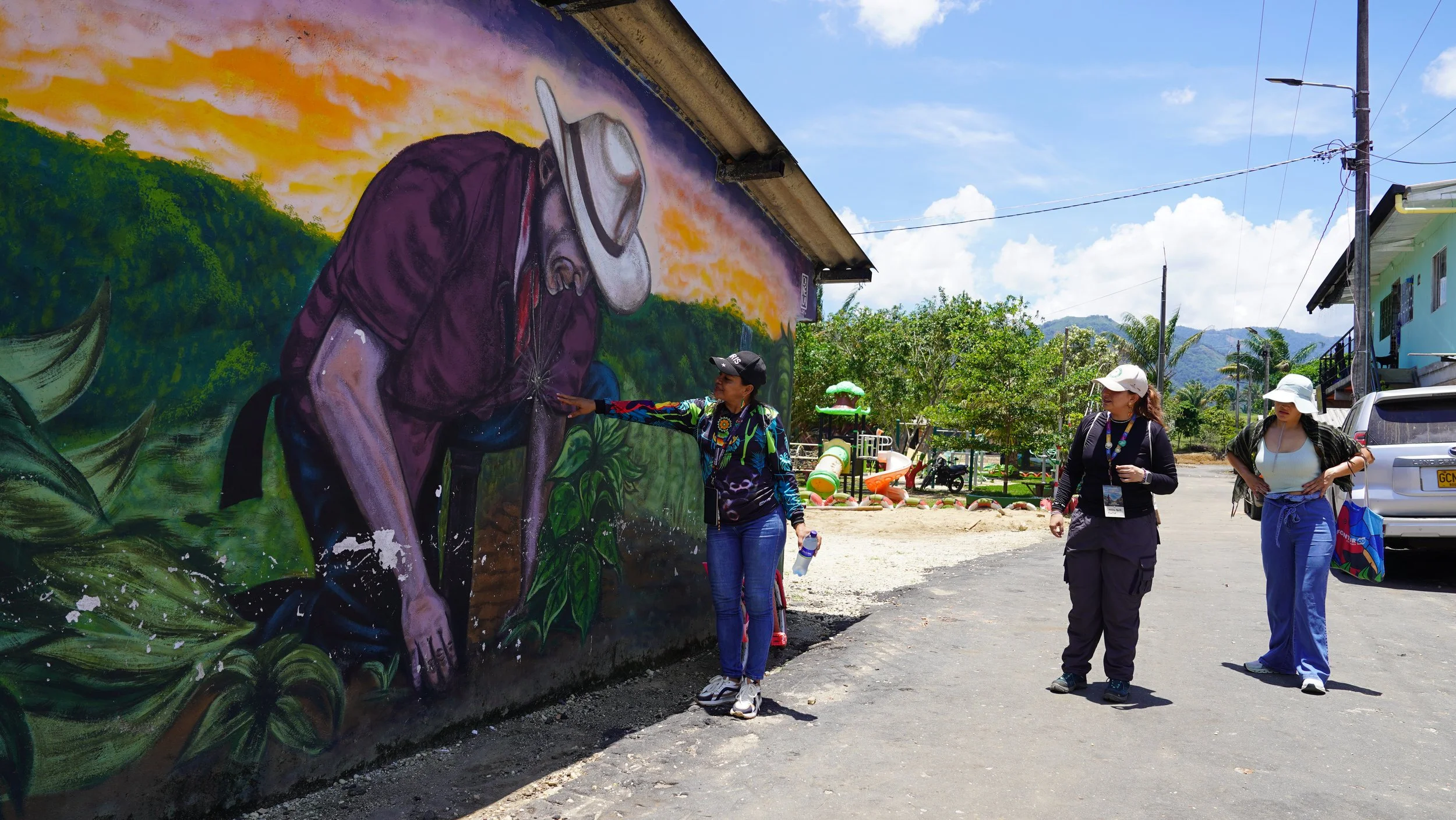 Mural de una persona con sombrero y lentes en un muro colorido, mientras tres mujeres observan y hablan frente a la pintura en un entorno al aire libre con casas, carros, y montañas al fondo.