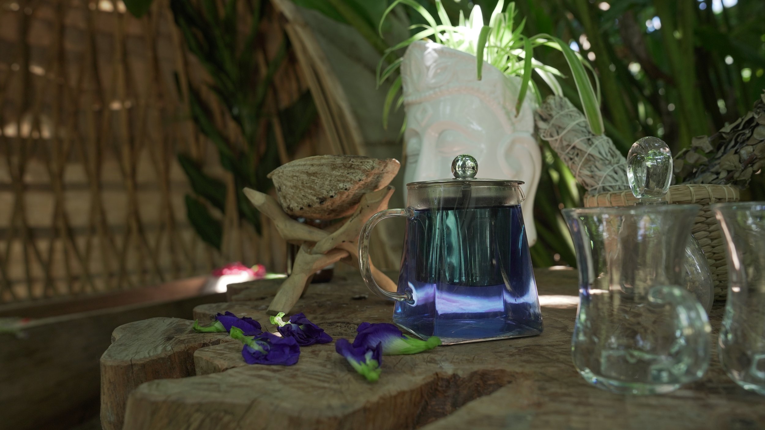Vaso de cristal con líquido azul, decorado con esfera de vidrio en la tapa, en un mesa de madera con flores moradas, y fondo de vegetación y pared de caña.