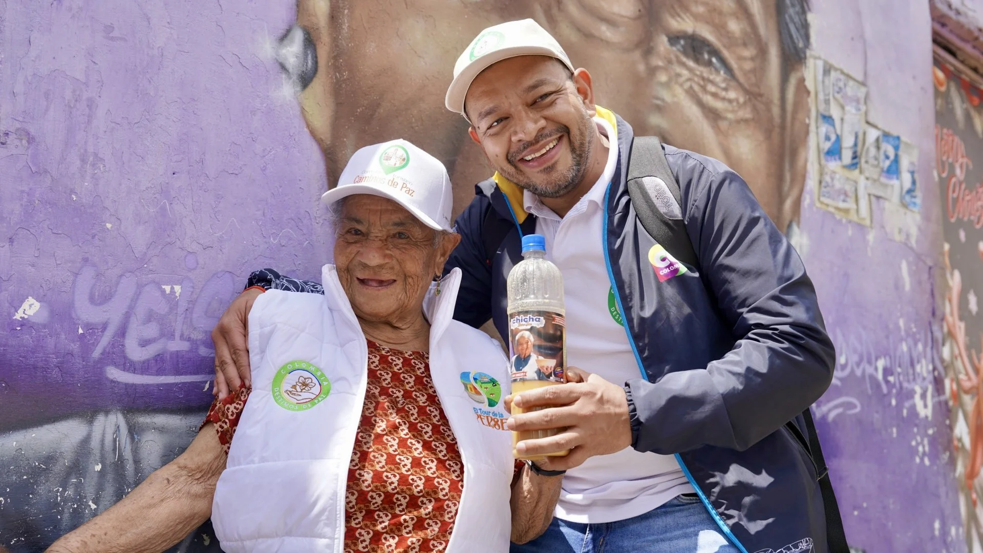 Un hombre y una mujer mayor sonrientes posando juntos en la calle, la mujer lleva una gorra blanca y una chaqueta con logotipos. La escena parece ser en un evento comunitario en Colombia, ya que ambos llevan insignias y gorros con el texto "Colombia Nos Une". La mujer sostiene un vaso decorado con una foto y texto, mientras que el fondo muestra un mural con una pared pintada de color púrpura y algunos dibujos pegados.