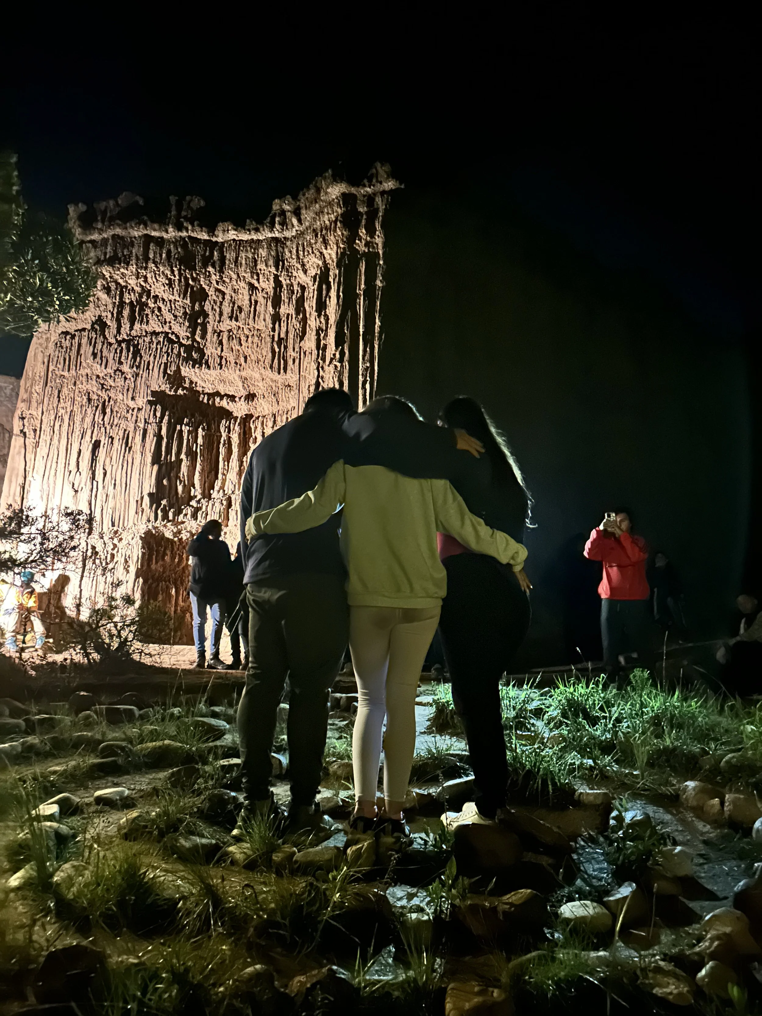 Grupo de personas en la noche, de espaldas, abrazadas, frente a una antigua estructura de piedra iluminada.