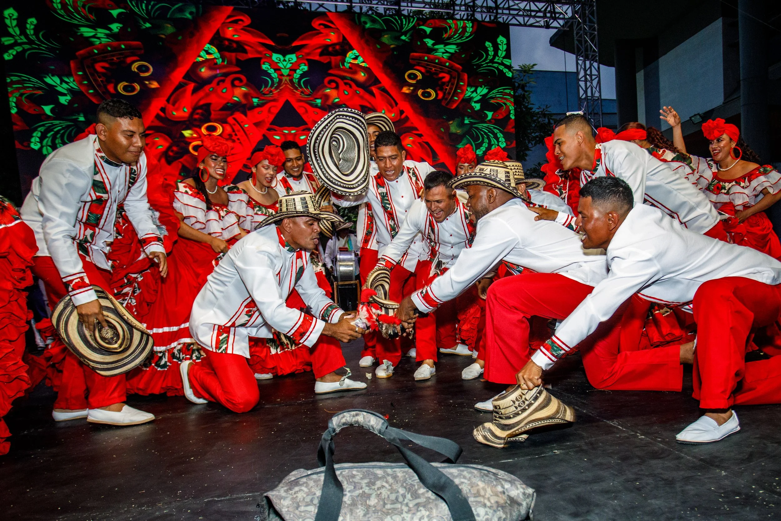 Grupo de personas bailando en trajes tradicionales en un escenario con fondo colorido y diseño artístico, durante una celebración cultural.