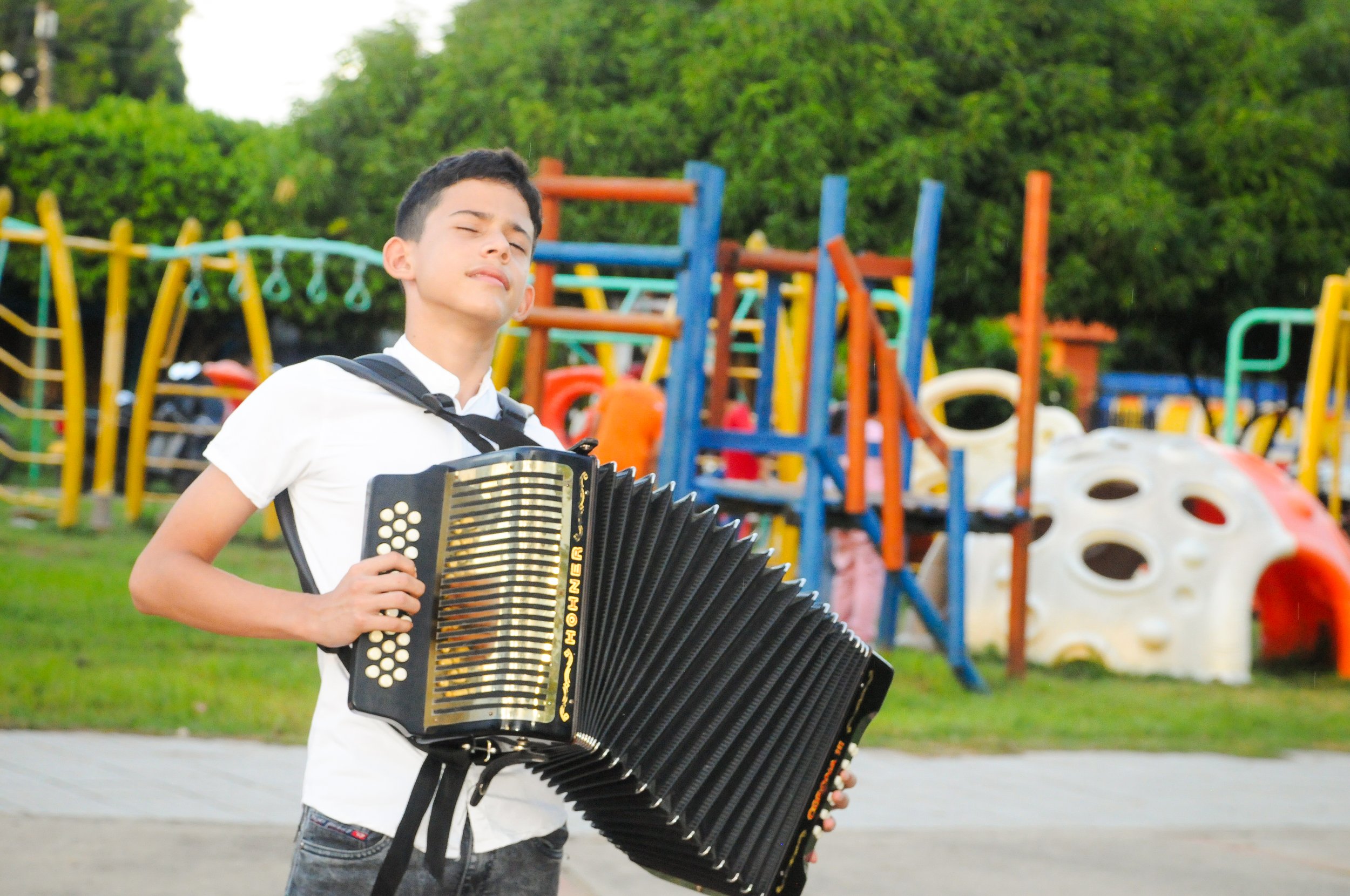 Joven tocando el acordeón en un parque con juegos infantiles de fondo.
