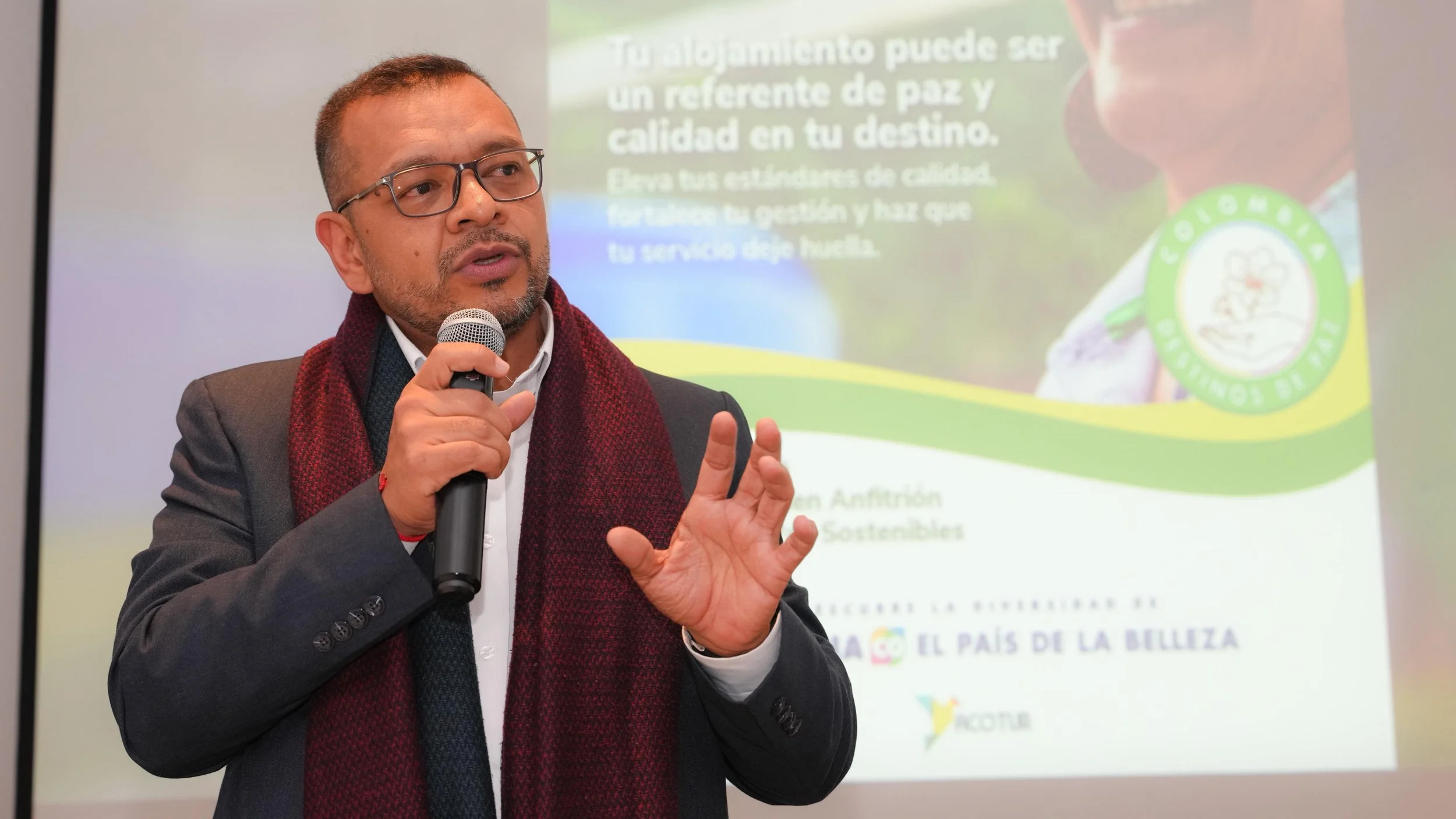 Hombre con gafas y barba, vestido con saco y bufanda, hablando con un micrófono en mano en una presentación, con una pantalla detrás que tiene texto en español sobre conciencia, paz y calidad en destinos turísticos.